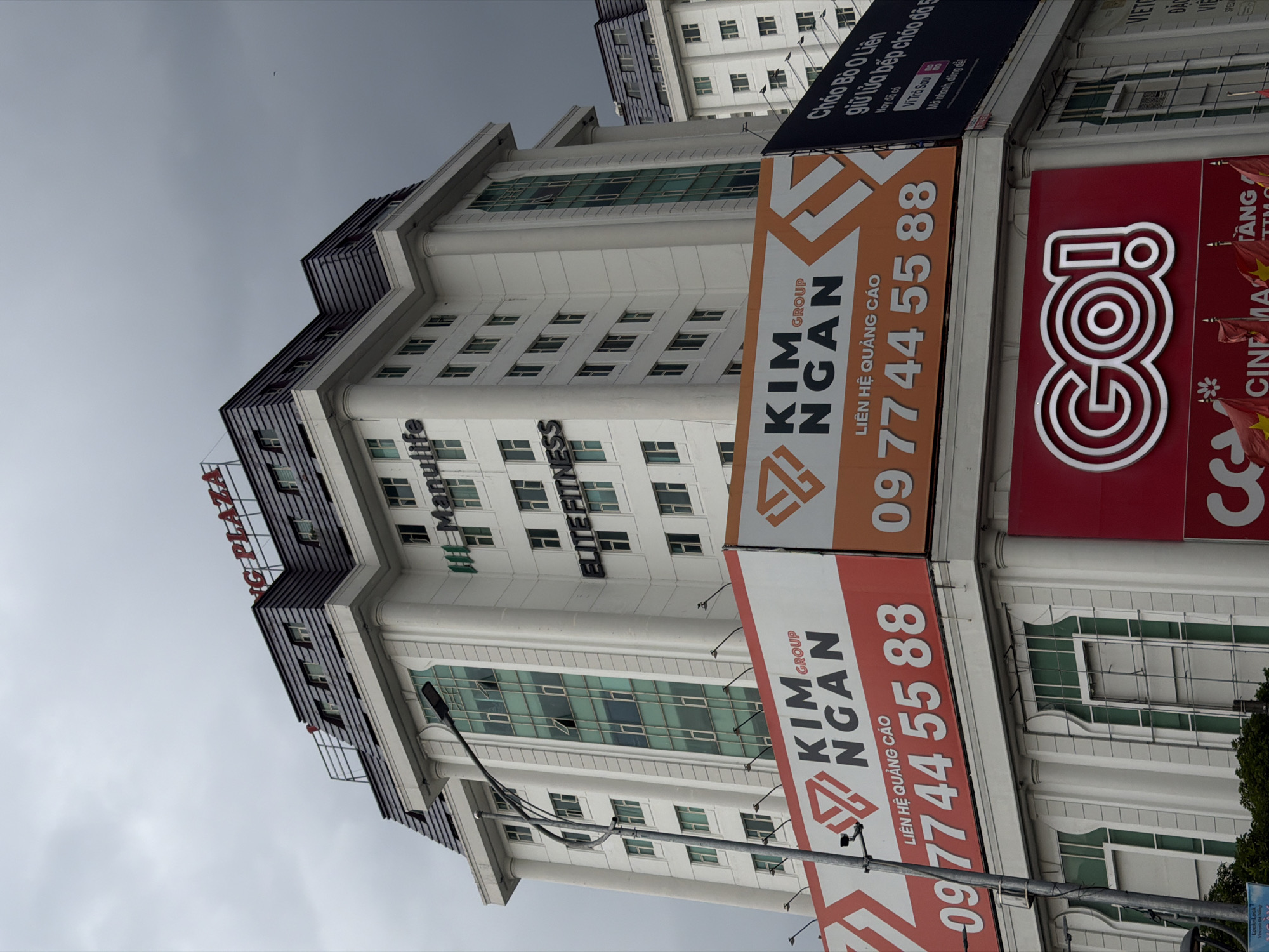 This is a photograph of a modern commercial building in Đà Nẵng, Vietnam. The building appears to be several stories high with a white facade and numerous windows. The image is taken from a low angle, looking upwards, and the building is slightly tilted.
Several signs are prominently displayed on the building's exterior. On the left, a red sign reads "NG PLAZA". Further down, a green sign with white text says "M.life" and "TNESS".
Dominating the right side of the image are two large vertical banners for "KIM GROUP NGAN". The banners are orange and white with large text and a phone number, "0977445588", repeated. Above these banners, a dark blue sign contains Vietnamese text, including "Chào Bô 0 Liên", "giữ lửa bếp chảo đã 5", "Nay đã có", "Vì Trà Sau", and "Mở nhanh, dùng dễ".
To the far right, a bright red sign with white stylized lettering spells out "GO!". Below this, a sign with "CINEMA TANG" is partially visible, along with Vietnamese flags.
The sky is overcast and grey, suggesting a cloudy or perhaps rainy day. The overall scene depicts a commercial district with various businesses advertised. The perspective and the presence of signs point to a bustling urban environment focused on commerce and entertainment. There are no people visible in the photograph.