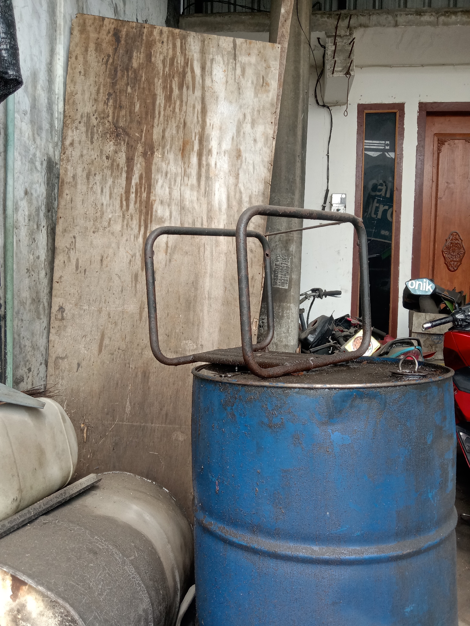 This is a medium shot of an outdoor storage area in Siwalan, Indonesia, with a focus on a blue metal barrel and a rusted metal frame on top. The barrel, approximately one meter in height, is weathered and appears to be a repurposed oil drum. It is situated on a dirt or concrete surface. Resting on the barrel is a rectangular metal frame with rounded corners, possibly part of a rack or stand.
In the background, to the left of the barrel, is a large, weathered piece of plywood leaning against a textured concrete wall. To the right of the barrel, a portion of a red motorcycle is visible, along with another, darker colored motorcycle behind it. Further back, a wooden door with intricate carvings is visible, suggesting a residential or commercial building. Wires and conduits are strung along the exterior of a white-walled structure next to the door. In the foreground, to the left of the blue barrel, are parts of two other large containers, one white and one dark gray, also appearing to be barrels or tanks. The overall scene depicts a cluttered and utilitarian outdoor space.
