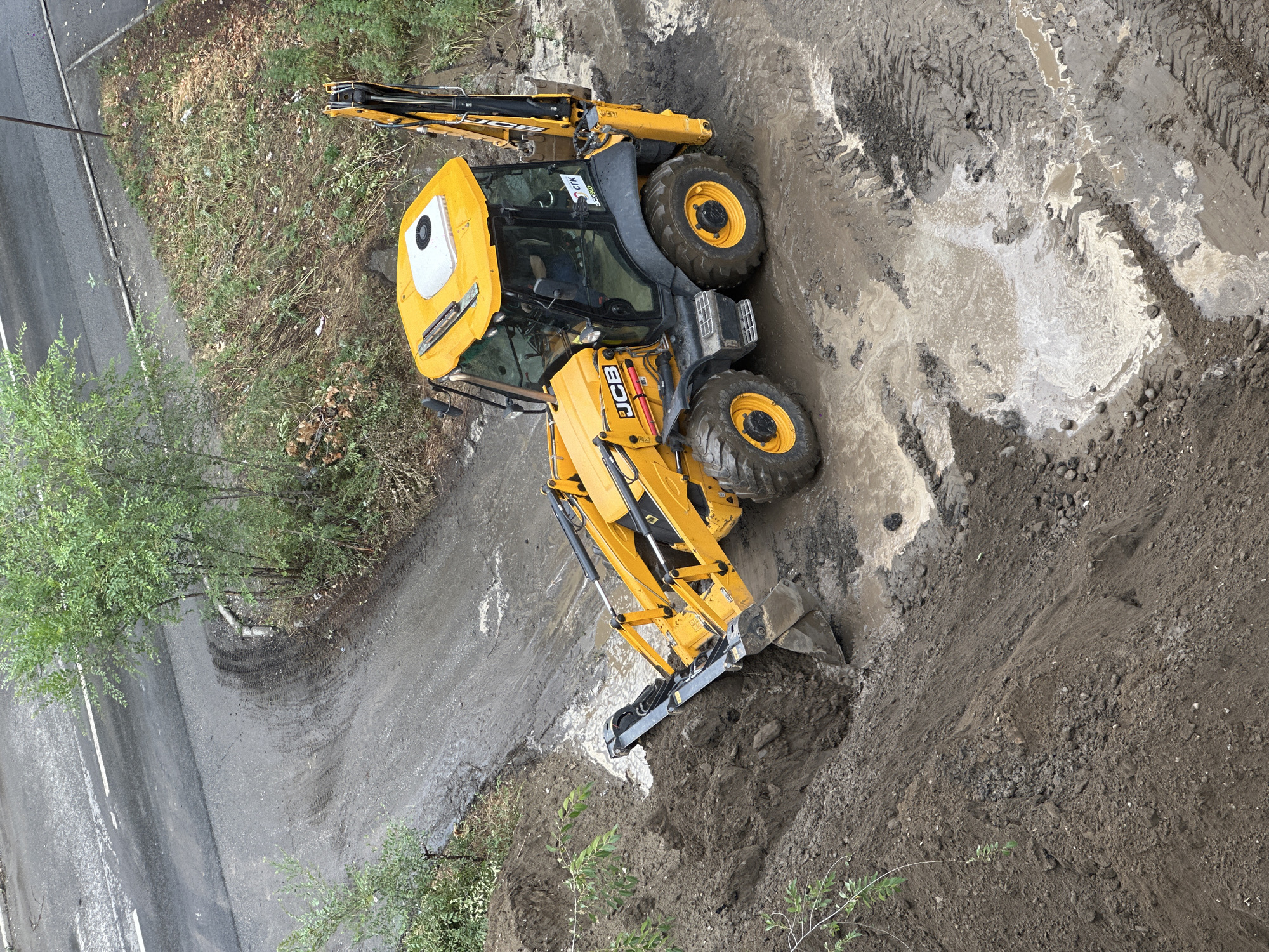 An aerial, high-angle view captures a yellow JCB backhoe loader operating in a muddy, excavated area adjacent to a paved road. The backhoe's front loader is actively digging into a pile of dark brown soil, which has been excavated from a trench visible in the foreground. The ground is saturated with water and mud, with visible tire tracks from the heavy machinery. Green foliage and trees are present on the right and left sides of the scene, bordering the construction area. The road in the background shows tire marks and some scattered debris. No people are visible in the image. The scene suggests ongoing construction or excavation work. The context of Biysk, Russia, is not visually represented in the image itself.
