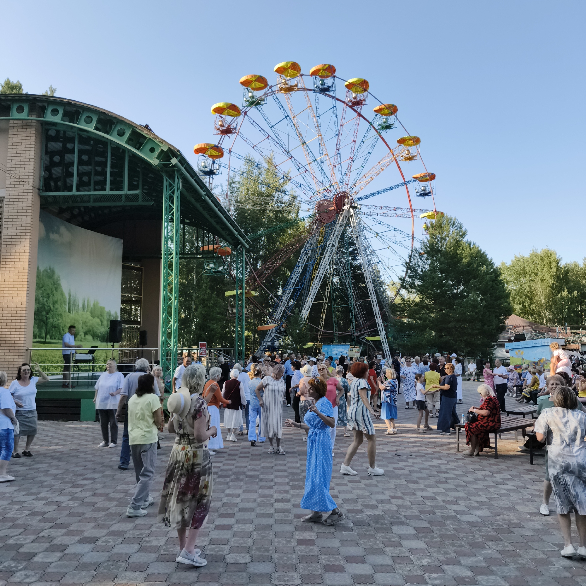 The image depicts a scene at an outdoor amusement park in Chaikovsky, Russia, with a Ferris wheel prominently featured in the background. A group of people are gathered, some appearing to be dancing or socializing in front of a stage area. The stage has a backdrop painted with a landscape of trees and sky. The ground is paved with a checkered pattern of reddish-brown and beige tiles. The overall atmosphere suggests a community event or festival.
