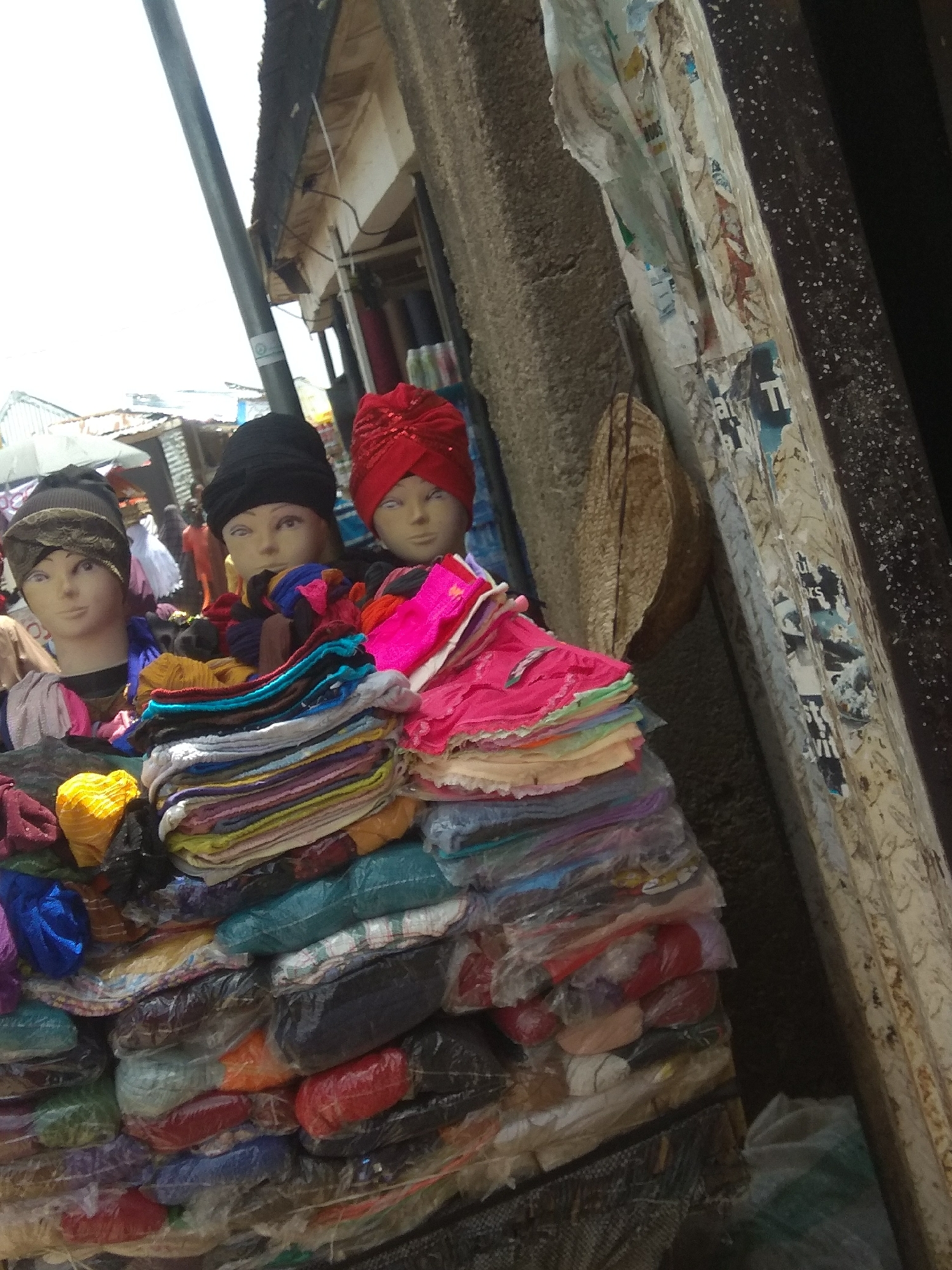 This image depicts a street market scene in Jalingo, Nigeria. The foreground is dominated by a vibrant display of textiles, primarily scarves and headwraps, stacked in neat piles. The fabrics come in a wide array of colors, including bright pinks, blues, yellows, and purples, with many appearing to be folded and wrapped in plastic.
In the background, three mannequin heads are visible, each adorned with a different headwrap. The left mannequin wears a dark, textured wrap. The middle mannequin sports a black turban, and the right one is prominently featured with a striking red sequined turban. Behind the mannequins and the textile display, glimpses of market stalls and structures can be seen, suggesting a bustling commercial area. A utility pole with some signage is also present on the left side of the frame. The overall atmosphere appears to be that of an active marketplace, likely during daylight hours, with the bright colors and outdoor setting indicative of such an environment. The texture of the building materials and the style of the headwear might offer clues to the specific cultural context of the location.