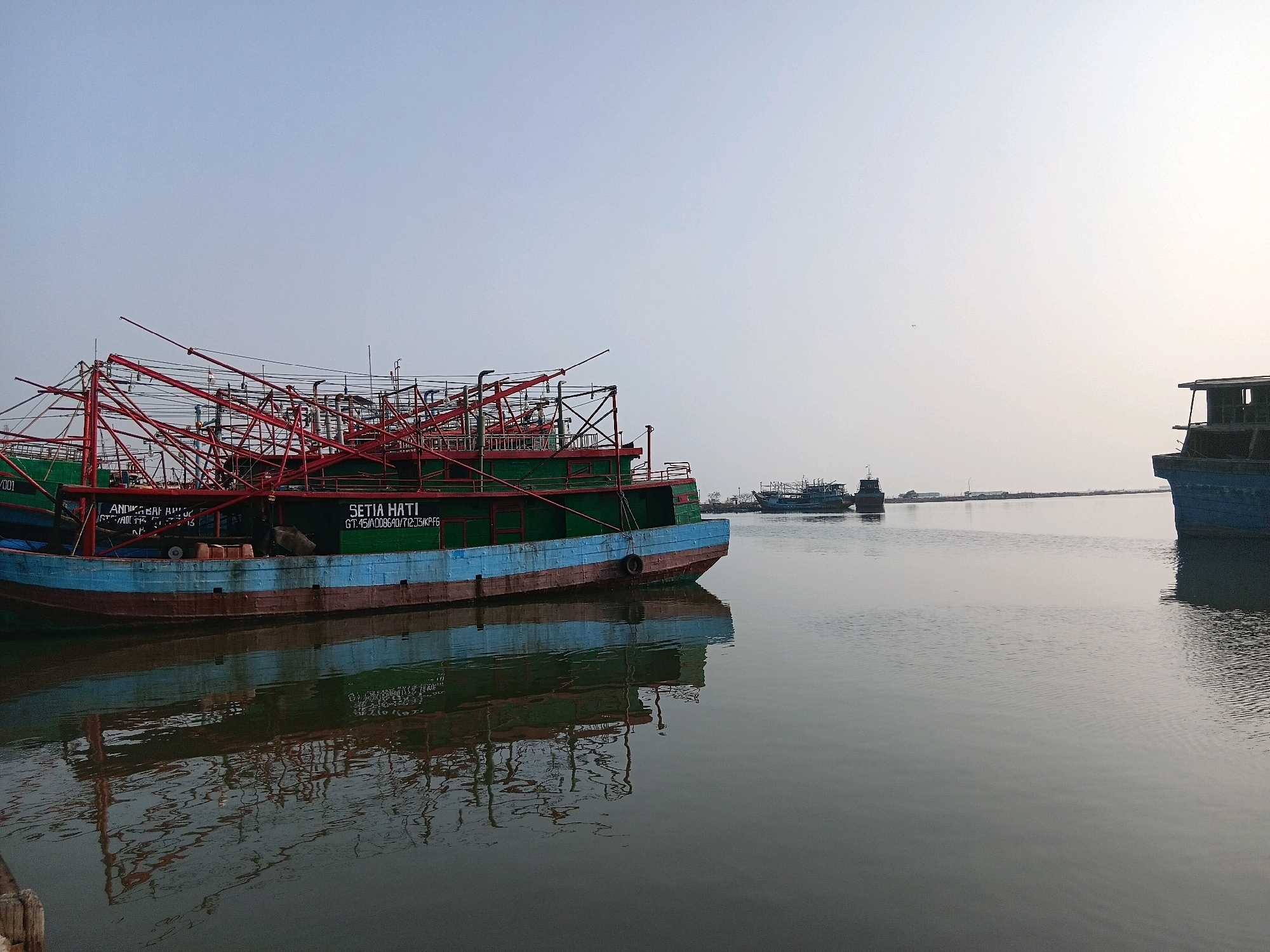 A fleet of green and blue fishing boats are docked on a calm body of water. The boats are lined up closely together, with the foreground boat dominating the left side of the frame. This boat is painted green and blue, with a rusty red hull. A complex network of red metal structures, likely fishing equipment or cranes, rises from the deck of the boat. Text on the side of the boat reads "SETIA HATI". The water reflects the boats and their equipment, creating a mirror image. In the background, other fishing boats are visible, as well as a hazy shoreline with some structures. The sky is a pale, overcast blue, with a slight gradient towards the horizon. The scene appears to be a harbor or fishing port, likely located in Cirebon, Indonesia, given the style of the boats and the presence of typical fishing vessels common in Indonesian waters. There are no people visible in the image.