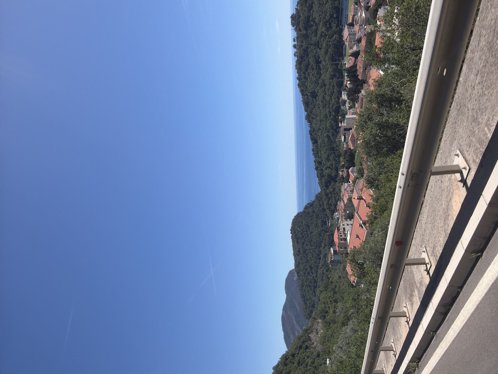 The image captures a scenic view of Novoselje, Montenegro, on a clear day. In the foreground, a metal guardrail runs horizontally across the bottom of the frame, separating a paved road from a vegetated slope. The road itself has a white line marking its edge. Beyond the guardrail, a cluster of buildings with red-tiled roofs are visible, nestled amongst lush green trees. The town appears to be situated on a hillside, with more greenery extending up the slopes. In the middle ground, rolling hills covered in dense forest lead towards the sea. The ocean is a deep blue, meeting the horizon under a vast, clear blue sky. A faint contrail is visible in the sky. The overall scene depicts a coastal town surrounded by natural beauty.