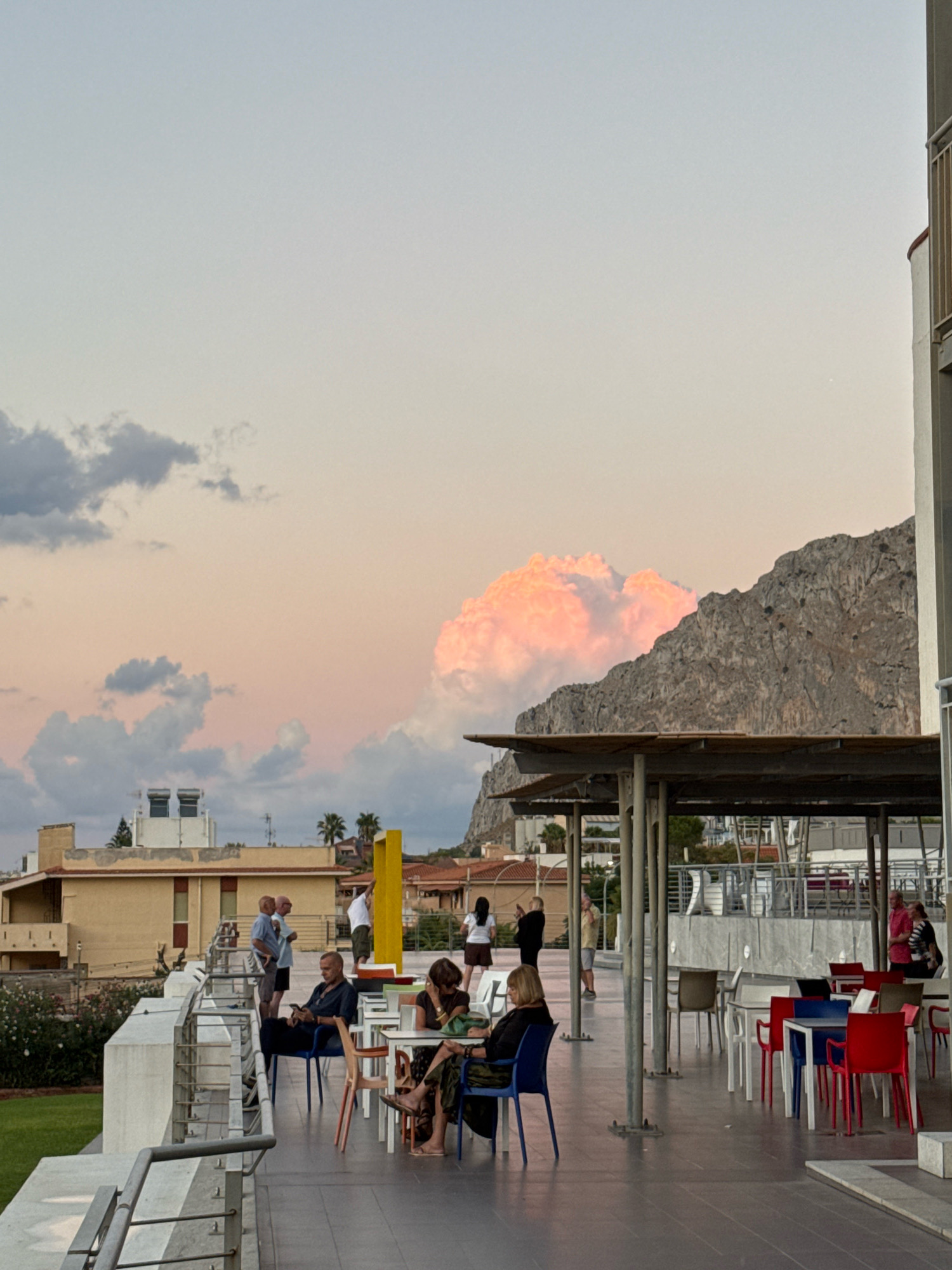 The image depicts a rooftop terrace area with people seated at tables and standing in groups. In the background, a large mountain looms over a town with yellow buildings and scattered palm trees. The sky is partly cloudy with a soft, pastel glow, suggesting either sunrise or sunset.
On the terrace, several tables with chairs are arranged. Some tables are empty, while others are occupied by individuals and small groups. A man in a dark shirt is seated alone, looking at his phone. A woman in a patterned skirt is seated at another table, with her hand near her face. Further back, a group of people are standing and talking near a yellow pillar. Another group, including a man in a pink and white striped shirt, are gathered near red chairs at a table.
The architecture of the terrace features a modern, open-air design with a sloped roof supported by pillars. A white railing runs along the edge of the terrace, overlooking a green lawn below. The overall atmosphere appears relaxed and casual, with people enjoying the outdoor space.
The scene is situated in Capaci, Italy, as indicated by the distinctive mountain in the background, which is likely Monte Pellegrino, a prominent landmark visible from the town. The presence of palm trees and the Mediterranean architectural style of the buildings further suggest this location. The lighting and cloud formations indicate a time of day when natural light is soft and diffused, creating a serene ambiance.