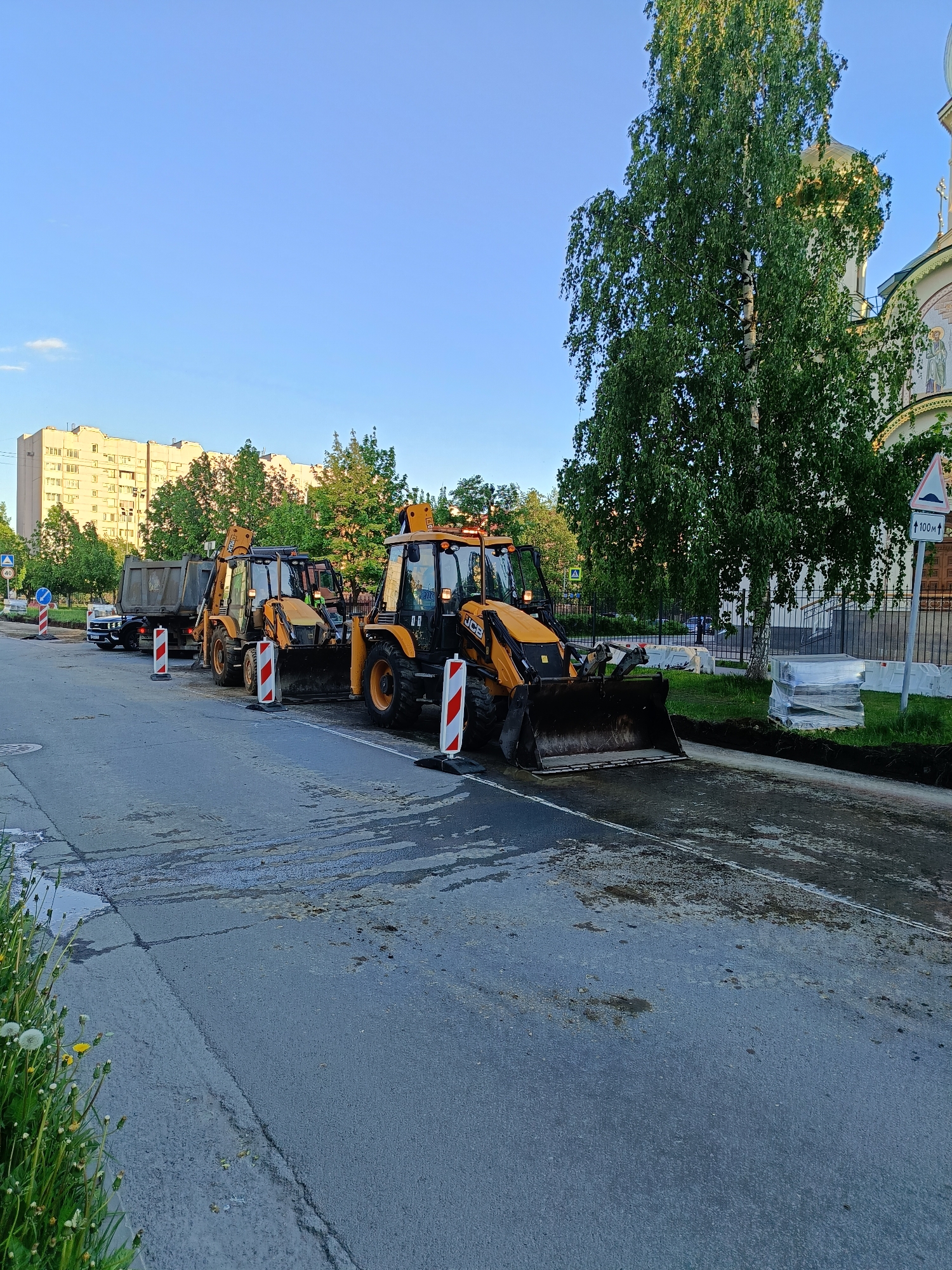 The image shows a street scene in a city, possibly in Russia, with construction vehicles and workers repairing the road.
Two yellow JCB backhoe loaders are parked on a section of the road marked with red and white striped barriers. Behind them, a dump truck is present. The road surface appears to be wet or partially cleaned.
In the background, there's a building with multiple floors, likely an apartment building. To the right, a large tree partially obscures a building with a golden dome, possibly a church or cathedral. A road sign indicating a bump is visible.
Construction work is underway, and the location appears to be an urban setting.