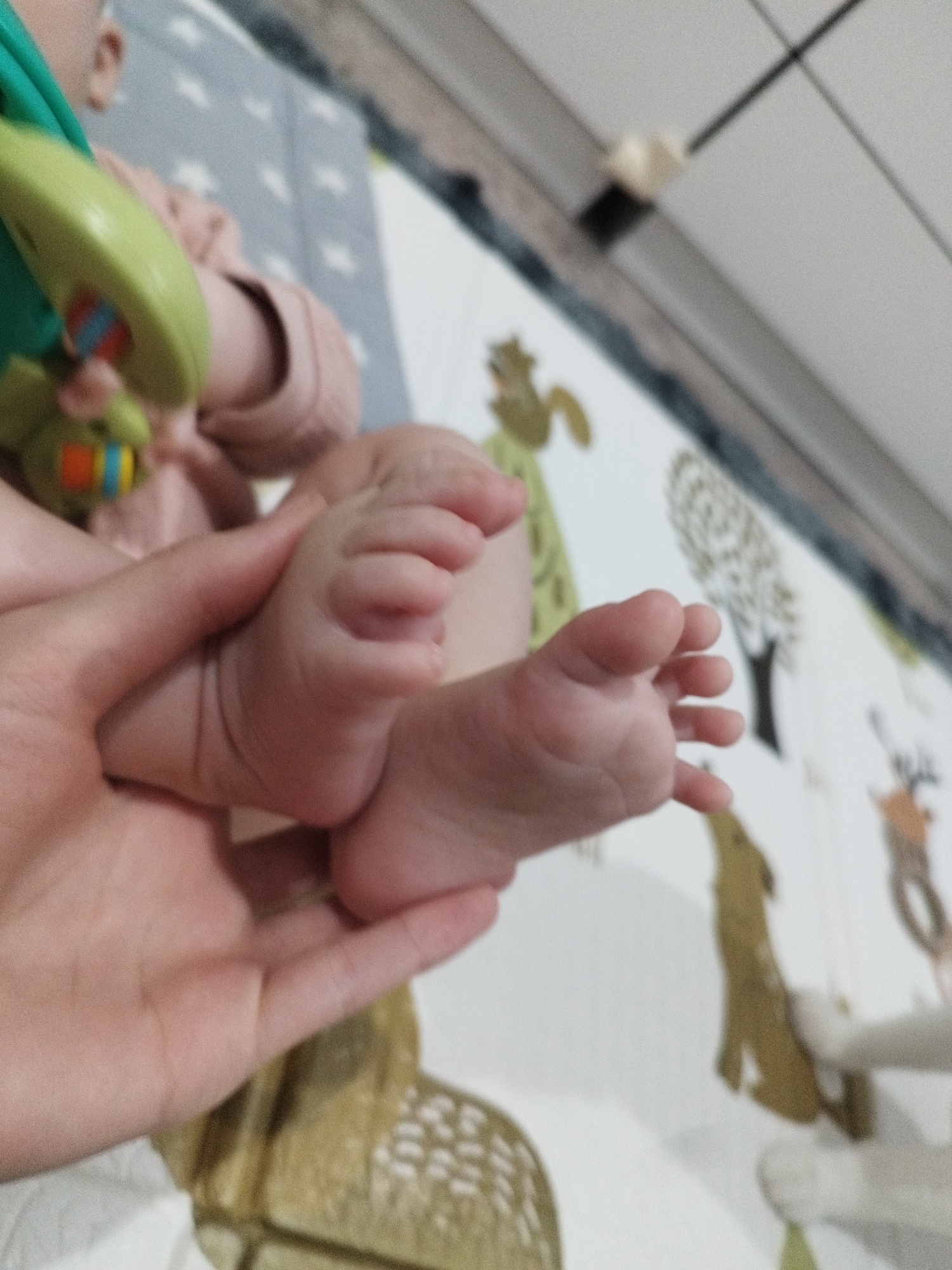A close-up shot focuses on a pair of infant feet being held in an adult hand. The background is a play mat with cartoon animal illustrations, including deer, trees, and a squirrel-like creature. The location is impossible to determine from the content of the image alone.
