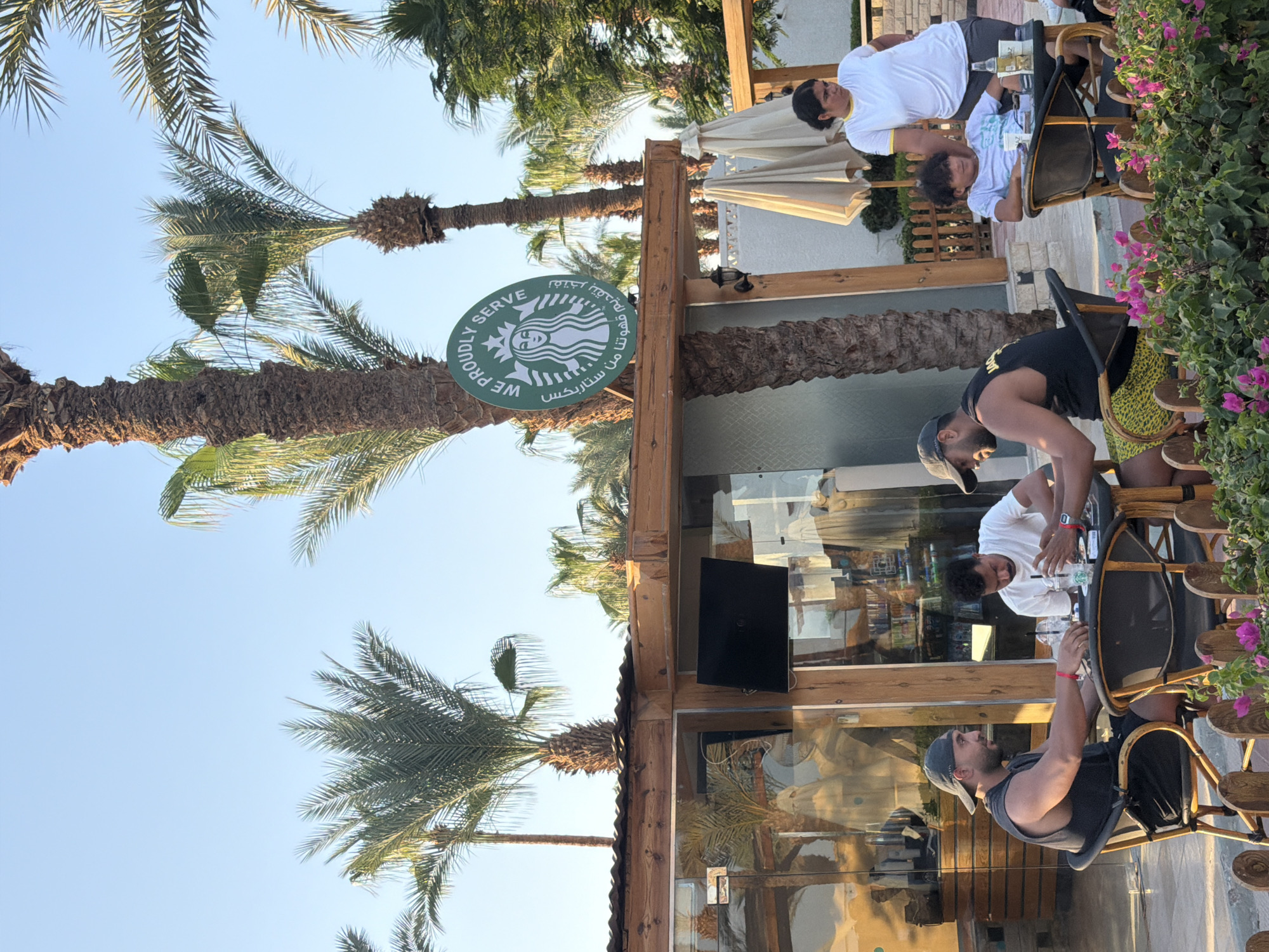 A medium shot captures a Starbucks cafe in Sharm El Sheikh, Egypt, on a clear day. Palm trees frame the scene, with their fronds silhouetted against a pale blue sky. The Starbucks logo is prominently displayed on a green circular sign above the cafe's wooden and glass facade.
Several individuals are seated at tables outside the cafe. In the foreground left, a man wearing a black tank top and a baseball cap is seated at a table, looking to his left. He appears to be interacting with someone off-camera or examining something on the table. Another man in a white t-shirt is seated across from him, looking down at the table. To the right, a young person in a white t-shirt is seated, looking towards the camera. A woman in a white t-shirt with yellow trim stands behind the table with the young person, her body angled away from the camera.
The cafe itself has a rustic wooden structure with large glass windows, offering a glimpse of the interior. Several tables and chairs are arranged outside. In the background, beach umbrellas are visible, suggesting a resort or beachside location. The overall atmosphere is casual and relaxed, indicative of a cafe in a tourist destination. The presence of Starbucks indicates a modern, branded establishment within the Egyptian city.