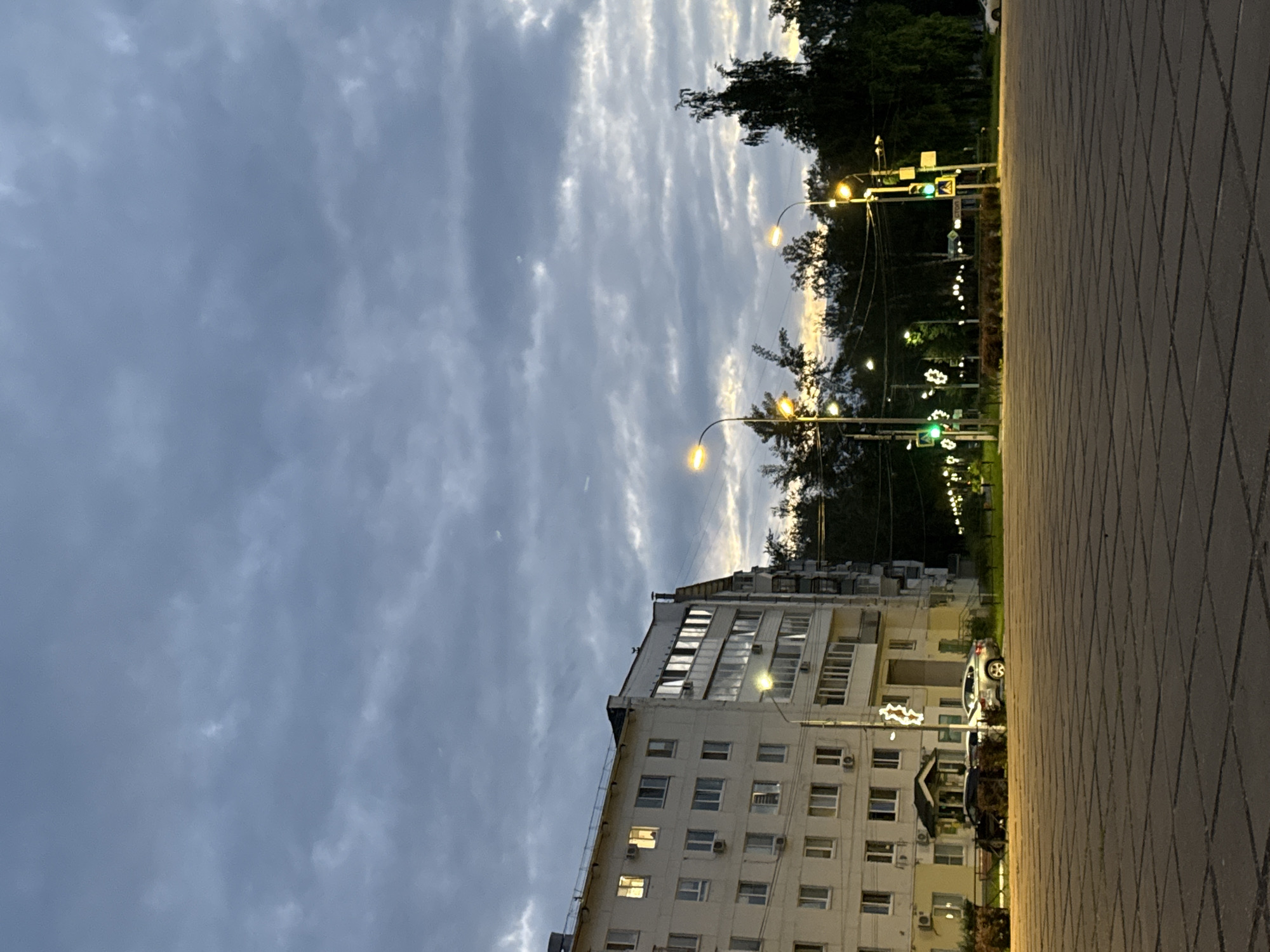 The image depicts a street scene in the city of Chaikovsky, Russia, during twilight. On the left is a multi-story apartment building with cream-colored facade and numerous windows, some of which are illuminated from within. In front of the building, several cars are parked. The foreground is a paved area with large rectangular stone tiles. A street with traffic lights, street lamps, and trees runs horizontally across the middle ground. The sky above is overcast with dense, dark clouds. The overall atmosphere suggests an urban environment at the end of the day, with artificial lights beginning to illuminate the scene against the fading natural light.