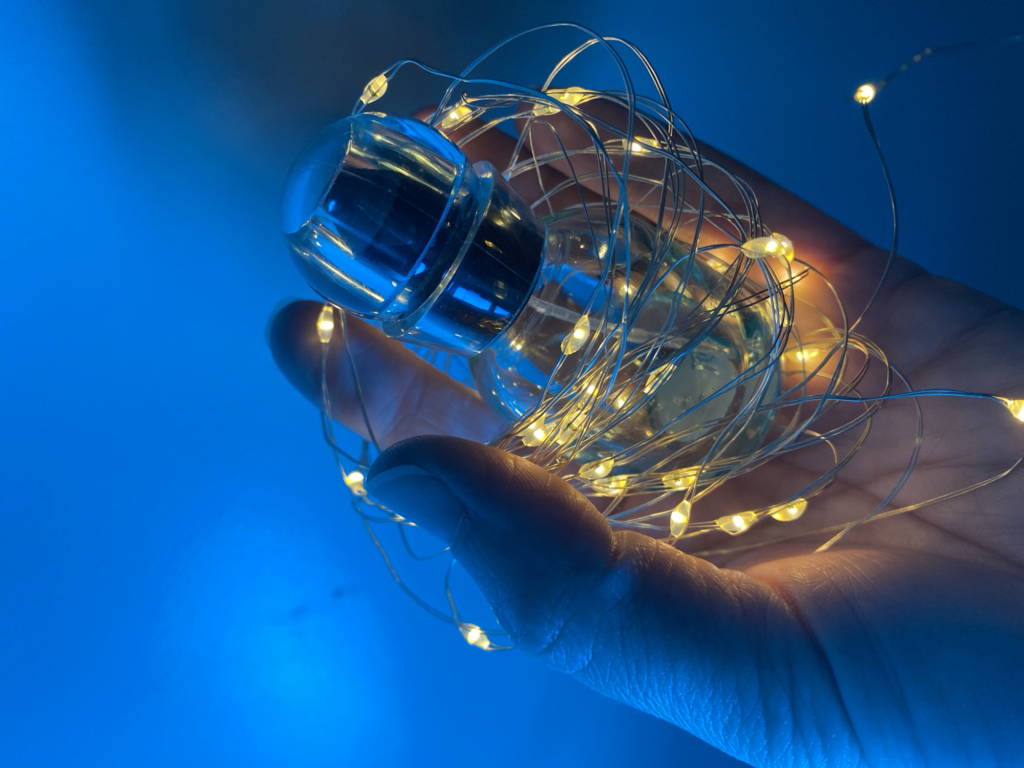 A hand holds a clear glass bottle wrapped in a string of small, warm-toned LED lights. The bottle has a cylindrical body and a darker-colored, potentially metallic, cap. The backdrop is a smooth, predominantly blue gradient. The lighting suggests a controlled indoor setting. There is no information available to infer the location is the city of Chaikovsky, Russia.