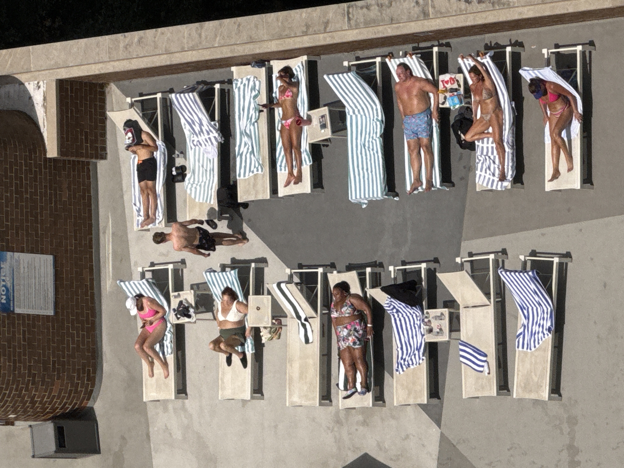 The image shows an aerial view of people sunbathing on loungers near a brick building. In the upper left corner, a trash can is visible next to the brick wall. A "NOTICE" sign is mounted on the brick wall.
There are at least seven rows of loungers, each with one or two people sunbathing. The loungers are arranged in a linear fashion. Some loungers have striped towels draped over them. Several people are wearing bikinis or swimwear, and some are wearing hats. One person is lying with a towel over their face. Another person appears to be looking at their phone. One man is walking between the loungers.
The ground appears to be a paved surface. In the background, there is greenery, suggesting an outdoor setting, possibly a hotel or resort. The lighting indicates a sunny day. The scene is observed from a high vantage point, looking downwards. The image is a static capture of people engaged in leisure activities.