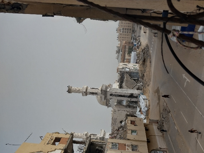 The image depicts a street-level scene in Khan Yunis, Palestinian Territory, characterized by widespread structural destruction under an overcast sky. In the center, a mosque with a prominent, largely intact minaret stands amidst significant rubble, its main building exhibiting extensive damage and exposed interior structures. To the left, a multi-story building, likely residential or commercial, shows severe damage with entire sections of walls collapsed, exposing rooms and rebar. Another damaged structure partially frames the scene on the far right.
On the asphalt road, a young individual is visible playing with a ball in the lower left foreground. Farther down the road, a solitary figure in dark clothing walks. In the mid-ground, substantial piles of concrete and building debris are scattered across the intersection and adjacent areas. Further down the street, beyond the immediate destruction, several individuals are observed walking, and some less damaged multi-story buildings are visible in the background, though the immediate foreground indicates a high level of devastation. The scene includes various objects such as corrugated metal sheets among the debris and a decorative metal railing partially obscuring the view from the foreground right.