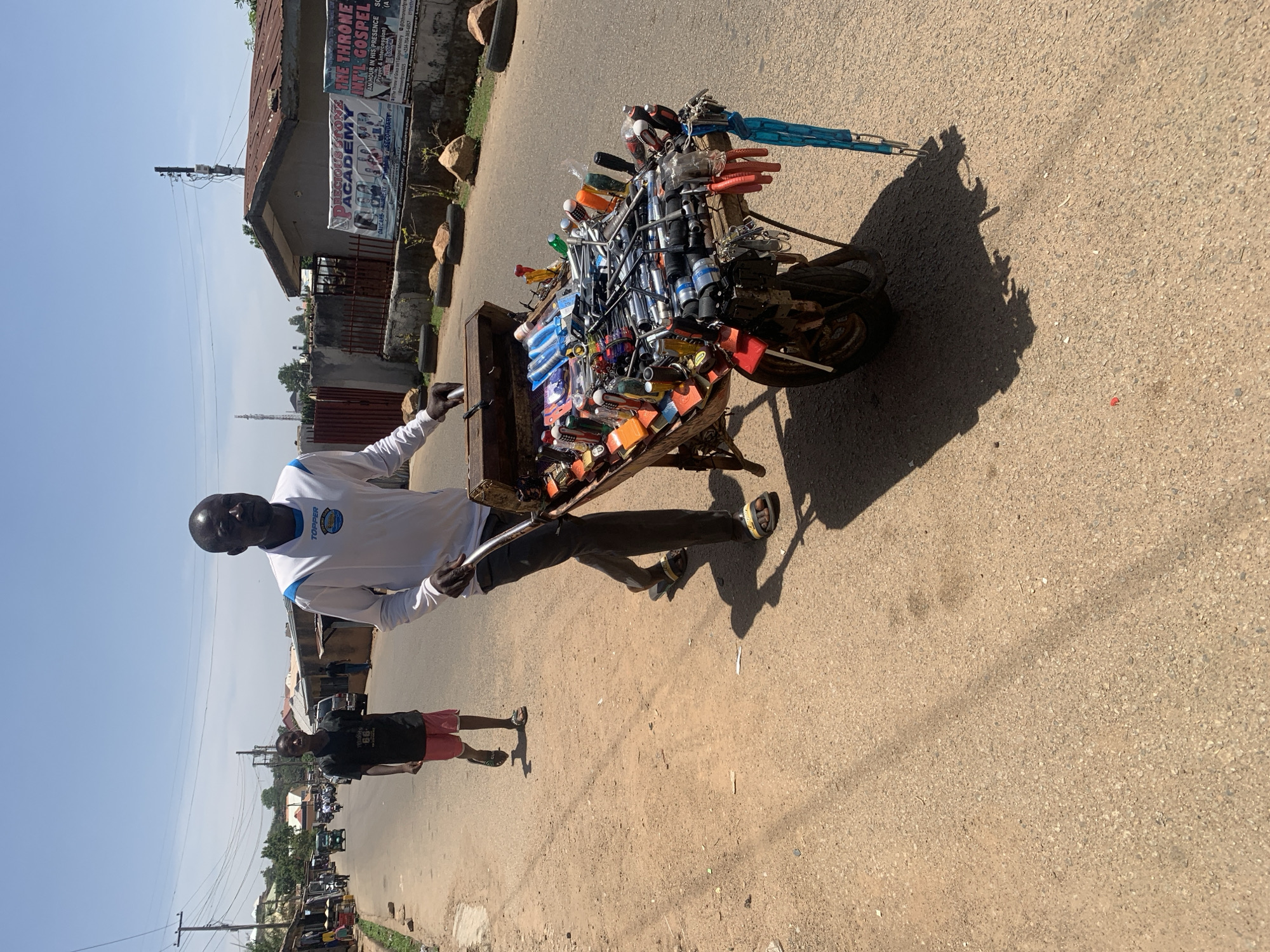 A street vendor in Jos, Nigeria, stands beside his heavily loaded mobile cart filled with various tools and hardware on a sunny day. The vendor, an African man in a white long-sleeved shirt with "TOPPER" visible on the chest and dark trousers, holds what appears to be a tool. His cart, likely attached to a bicycle (partially visible), is overflowing with a wide array of merchandise including screwdrivers, wrenches, sockets, pliers, and locks, meticulously arranged for display.
Behind the vendor, another African male, possibly a younger man or boy, in a dark t-shirt and red shorts, stands on the dusty paved road. The bright daylight casts distinct shadows, suggesting a time between mid-morning and early afternoon, under a clear blue sky.
The street scene features a row of older buildings with visible wear, one of which has a corrugated metal roof. Two banners are affixed to this building. One banner advertises "PRECIOUS STONE ACADEMY" offering DAYCARE, NURSERY, and SECONDARY education. The other banner promotes "THE THRONE INFL. GOSPEL" with a message about "AN HOUR IN HIS PRESENCE (Prayer & Intercession)" and contact details, mentioning its location "Adjacent ITF Corpers' Lodge." The image captures a snapshot of daily life and street commerce in an urban setting in Jos, Nigeria.