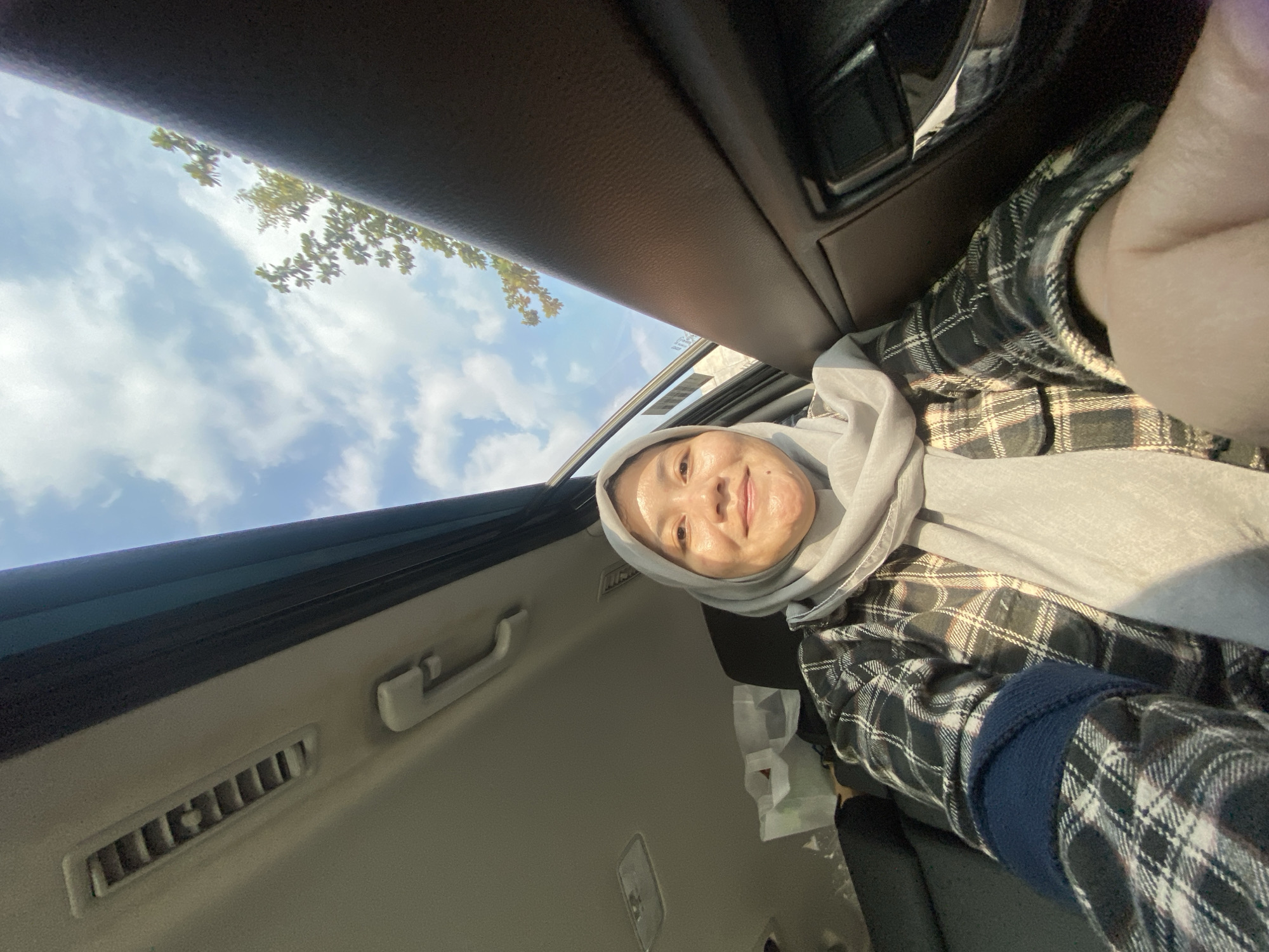 A young Indonesian woman wearing a light gray hijab and a black and white plaid shirt is taking a selfie inside a car. She is smiling and looking at the camera. The car appears to be in motion, as evidenced by the blurred trees and sky visible through the open sunroof and window. The interior of the car includes a black leather seat, a vent on the ceiling, and a door handle. The scene suggests the woman is traveling, possibly on a road trip or during her daily commute in Mandar Sari, Indonesia.