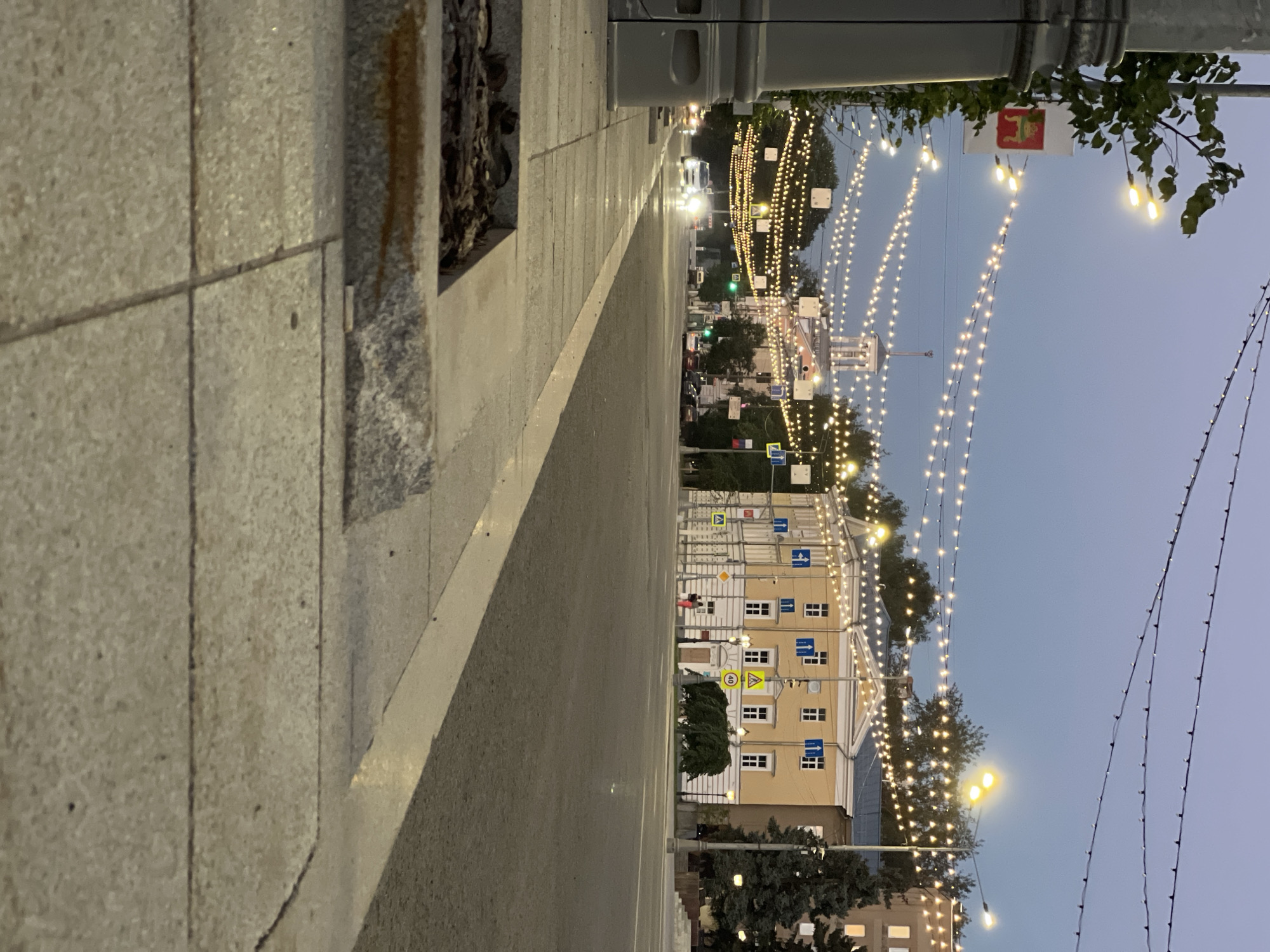 The image shows a street scene in Tver, Russia, likely during twilight or early evening. The foreground is dominated by a sidewalk made of large, light-colored rectangular paving stones, with a planter box containing dark mulch and some dry leaves. A gray, cylindrical lamppost or bollard is partially visible on the left.
Beyond the sidewalk is a paved road with visible lane markings. Several cars are driving on the road, some with headlights on. Across the street, a yellow building with white trim and numerous windows stands out. It appears to be a public building, possibly a government office or museum, given the signage and architectural style. A spire or tower-like structure is visible behind this building.
The street is illuminated by strings of decorative lights, hung in multiple parallel rows across the road. These lights are small and yellow, creating a festive atmosphere. Trees line the sides of the street, and more buildings are visible in the background, suggesting an urban environment. The sky is a dusky blue, indicating the time of day. There are a few pedestrians visible on the sidewalk in front of the yellow building, appearing as small figures in the distance. The overall impression is of a well-lit, pedestrian-friendly street in a Russian city.