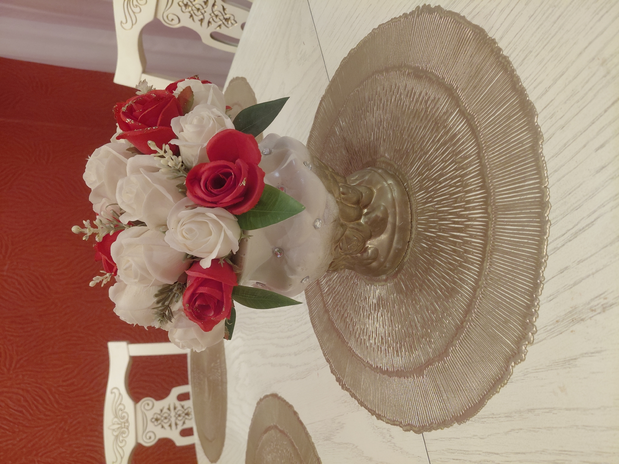 The image depicts a floral arrangement in a vase placed on a round, textured placemat on a wooden table. The arrangement consists of red and white roses, some with glitter accents. The vase is ornate and appears to be made of a metallic material, possibly gold, with some embellishments. Two white chairs with decorative backs are visible in the background, along with a textured red wall. The scene suggests an indoor setting, likely a dining area. The provided information states the location is the city of Malgobek, Russia.