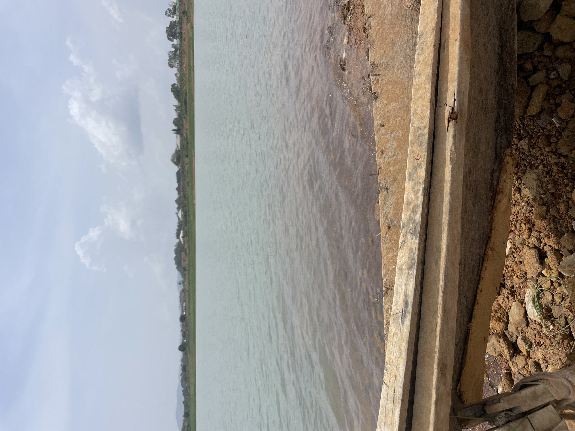 The image depicts a body of water in Jos, Nigeria, likely a dam or large pond, from an elevated perspective. A constructed ledge, possibly concrete or wood, runs along the right side of the frame. The water is a pale greenish-grey, reflecting the sky. The opposite bank features a strip of green vegetation and trees. The sky is overcast, with light clouds visible. The ground on the right of the ledge consists of dirt and small rocks.