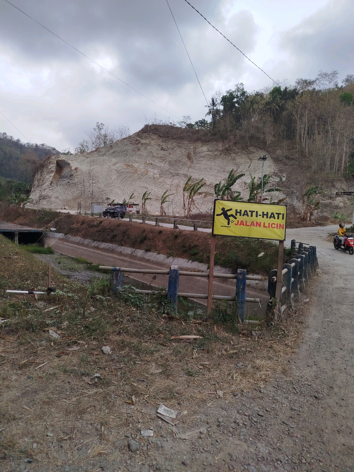 The image depicts a roadside scene in Kemangguhan, Indonesia. The environment is characterized by a steep, excavated hillside on the left, revealing layers of light-colored soil and rock. A narrow, concrete-lined canal runs parallel to the road. The road itself appears to be unpaved, consisting of gravel and dirt.
Several elements are present:
* **Signage:** A prominent yellow sign with black text and an icon of a person slipping is placed on the right side of the road. The text reads "HATI-HATI JALAN LICIN," which translates to "BEWARE SLIPPERY ROAD."
* **Vehicles:** A dark-colored SUV is parked on the road near some small structures. Further down the road, a person is riding a red scooter, wearing a yellow top.
* **Vegetation:** Sparse trees and dry grass are visible on the hillside and along the roadside. Banana trees with prominent leaves are also scattered throughout the scene.
* **Infrastructure:** Power lines are visible stretching across the cloudy sky. A solar-powered street light stands near the yellow sign. A fence with blue posts and grey railings runs along the edge of the road, separating it from the canal.
* **Atmosphere:** The sky is overcast, suggesting a potentially damp or humid climate. The overall impression is one of a rural or semi-rural area, possibly undergoing construction or land alteration, with a warning about road conditions.