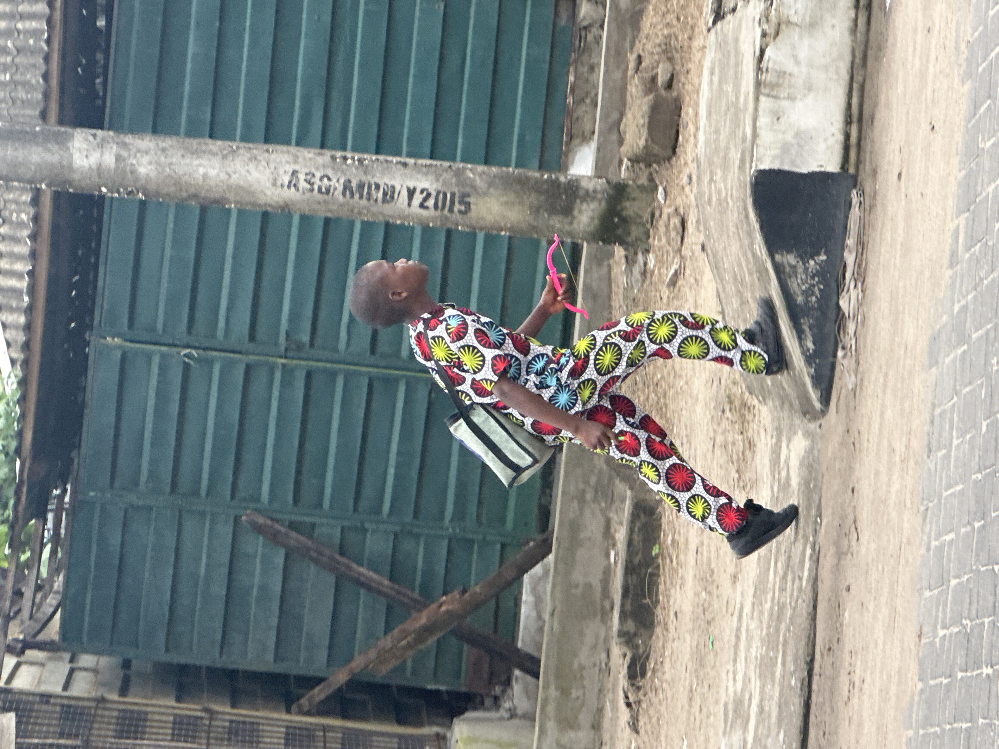 A young Black child, dressed in a vibrantly patterned outfit of white, red, yellow, and blue circles, is walking from left to right in the frame. The child carries a light-colored bag with a black strap over their left shoulder and holds a pink toy bow in their right hand. The child is wearing dark shoes and is on a sloped concrete path that transitions to a paved area. Behind the child is a tall, weathered concrete pole with some markings that are difficult to decipher, and a dark green corrugated metal gate. The surrounding environment appears to be an outdoor urban setting, with some debris and rough ground visible. The image seems to have been taken in Badagry, Nigeria, given the context of the image.