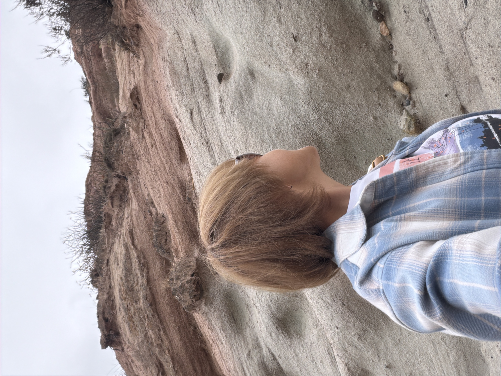 The image shows a person lying on a sandy, textured surface next to a rocky cliff. The person has blonde hair and is wearing a blue and white plaid shirt over a t-shirt with a nature-themed graphic. Sunglasses are visible on their head. The rocky cliff is reddish-brown and has some sparse vegetation growing from it. The sky appears overcast, suggesting a cloudy day. The scene is outdoors, with the sandy surface and rock formations indicating a natural, possibly coastal or desert environment. The person appears relaxed, looking upwards. There is no visible text in the image.