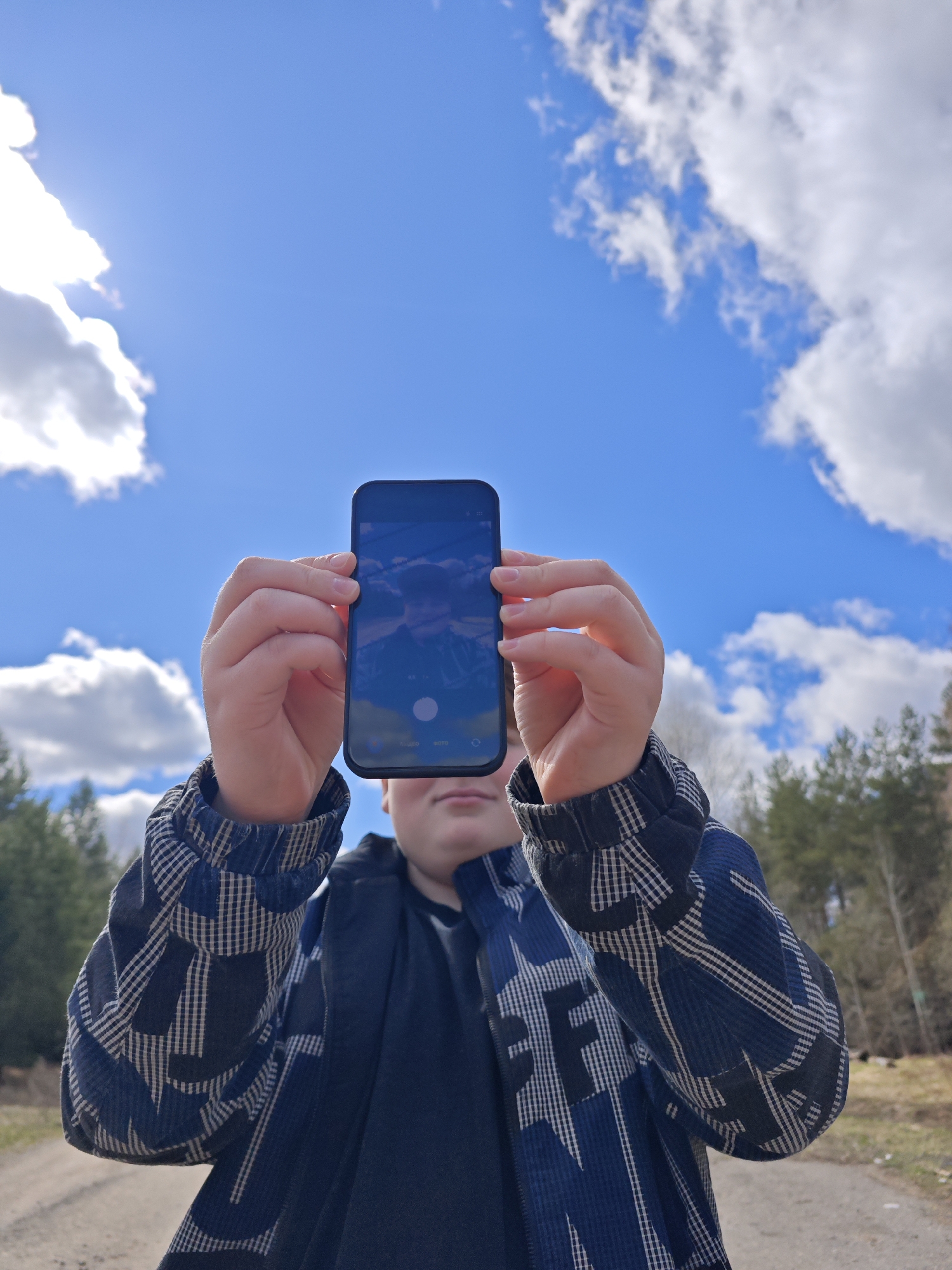 A person is holding a smartphone up towards a bright blue sky dotted with white clouds. The phone's screen displays a reflected image that includes a mountain peak. The person is wearing a navy blue jacket over a dark undershirt. The arms and hands holding the phone are visible. The background shows a dirt road leading towards a wooded area with various evergreen trees. The location is likely in the area of Chaikovsky, Russia, suggested by the style of clothing, landscape and reflected mountain.