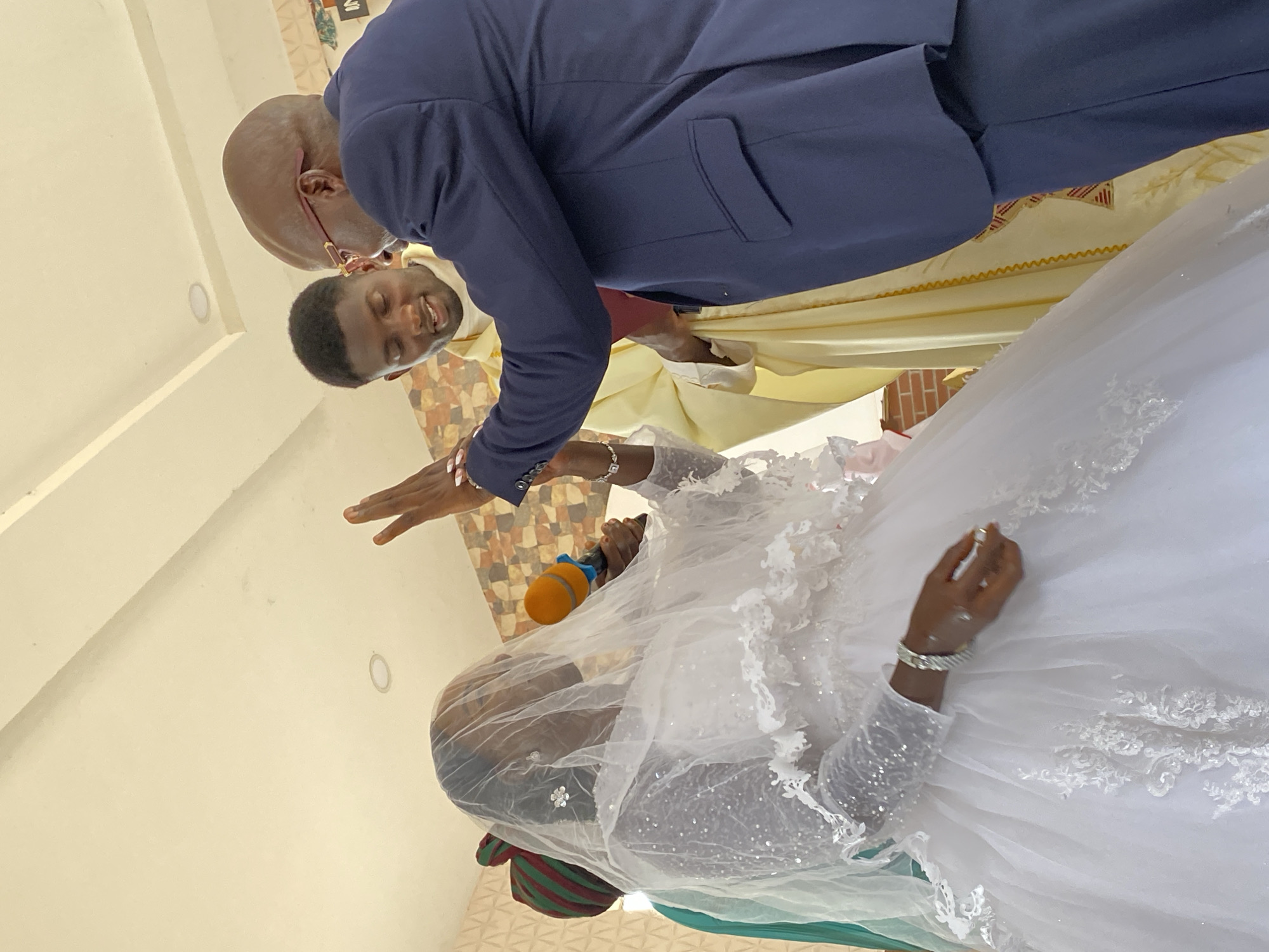 The image shows a wedding ceremony indoors in Kuru Dungus, Nigeria. A bride in a white gown and veil holds a microphone. Two men are above her: one in a navy suit and the other in a yellow robe. One of the men is making a gesture towards the bride with his hand.