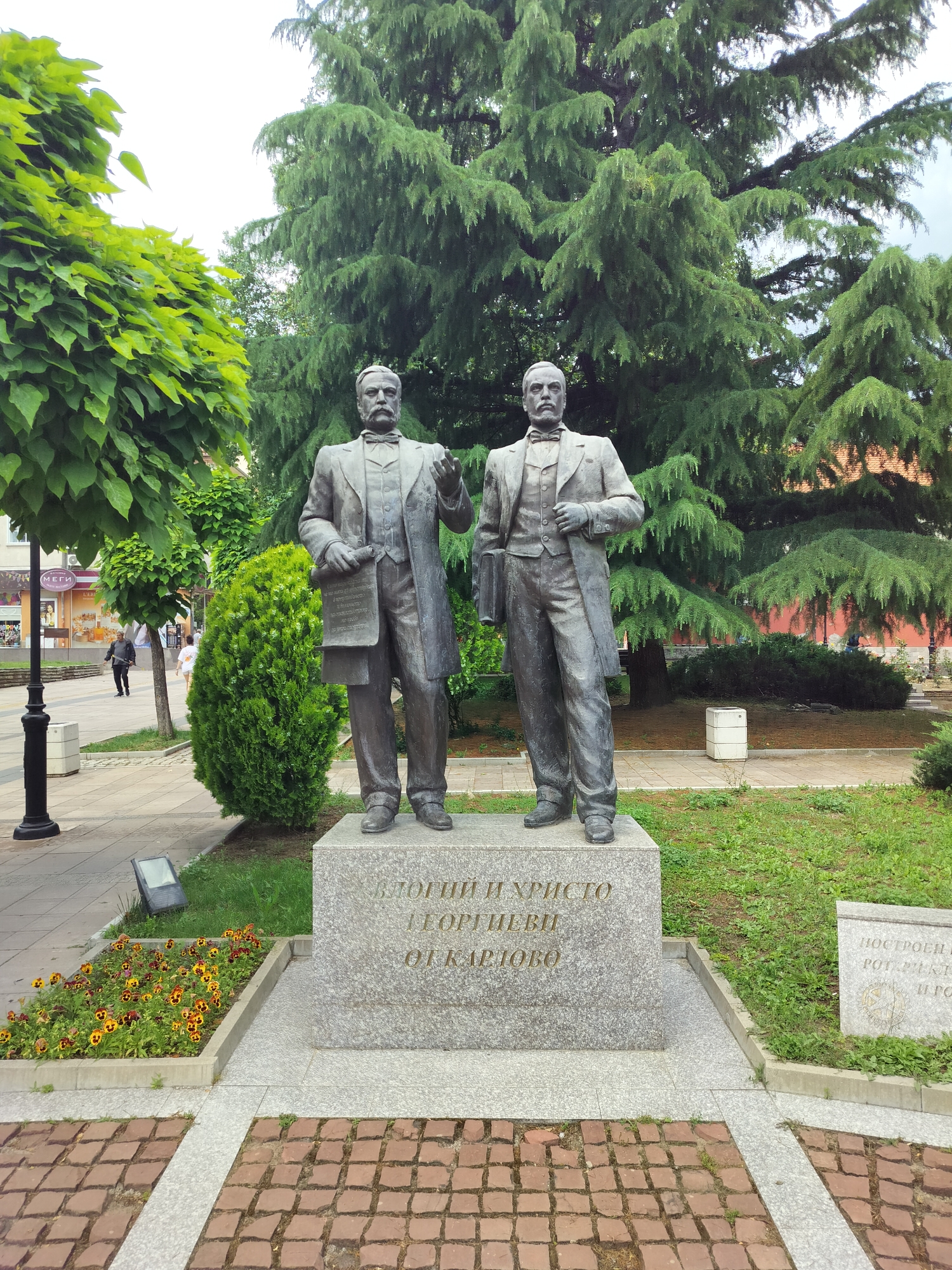 A bronze statue of two men stands in a public square in Karlovo, Bulgaria. The men, depicted in suits and ties, are positioned on a stone pedestal inscribed with Bulgarian text. The statue is set against a backdrop of lush green trees and a cloudy sky. In the foreground, a paved walkway is visible, bordered by flower beds containing colorful flowers. Other elements in the scene include a lamppost, a trash bin, and buildings in the distance. The overall atmosphere is serene and commemorative.