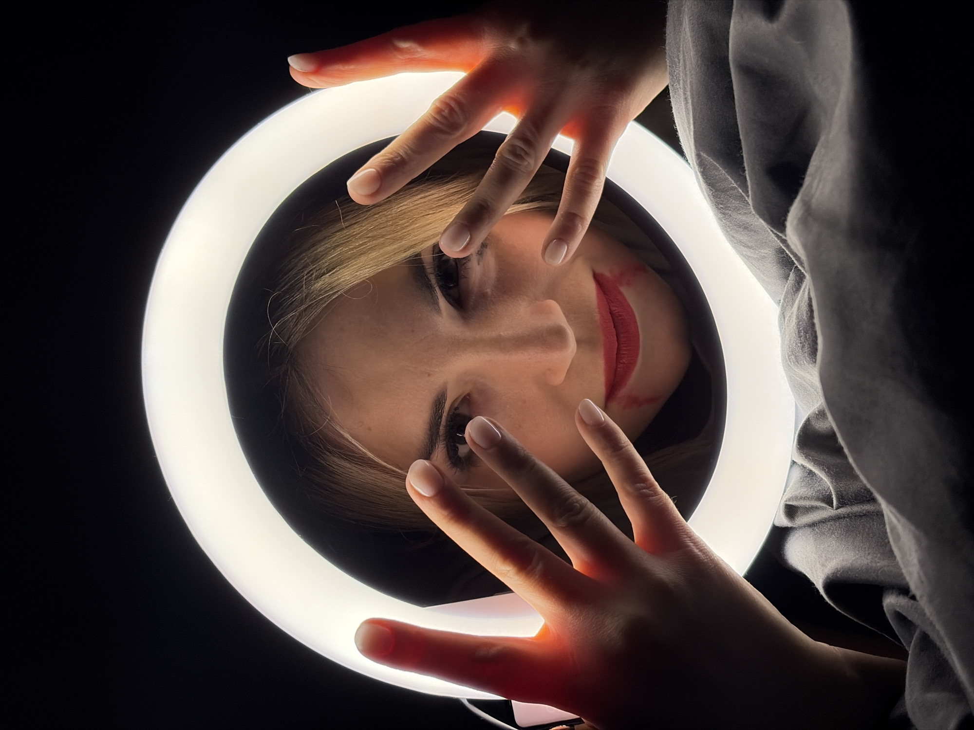 A medium shot shows a woman, facing down, with her face within a ring light. Her hands are above and below her face, partially obscuring her view. Her fingers have light pink polish. She has fair skin, brown eyes, and blonde hair. Her lips are painted red. Her mouth is slightly smeared to one side. She wears a dark hooded sweatshirt. The background is dark. The location is not identifiable as Budva, Montenegro.