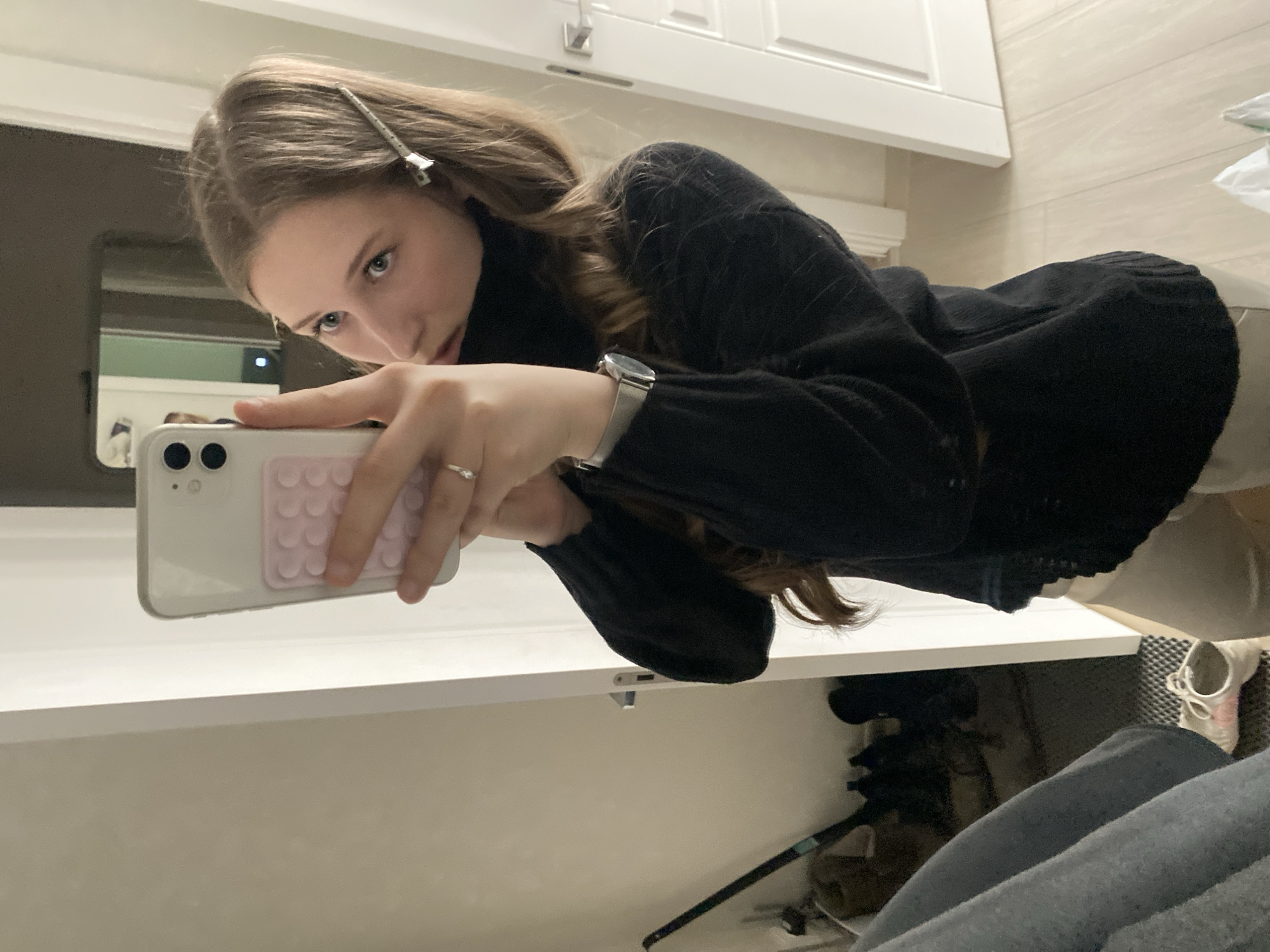 A young woman is taking a selfie in a bathroom. She is holding a white smartphone with a pink, textured grip on the back. She is wearing a black sweater and has a silver hair clip in her brown hair. She is also wearing a silver watch and a ring on her finger. The bathroom has white cabinets and light-colored tile. The presence of a mirror in the background indicates a bathroom setting. The location cannot be determined from the image.