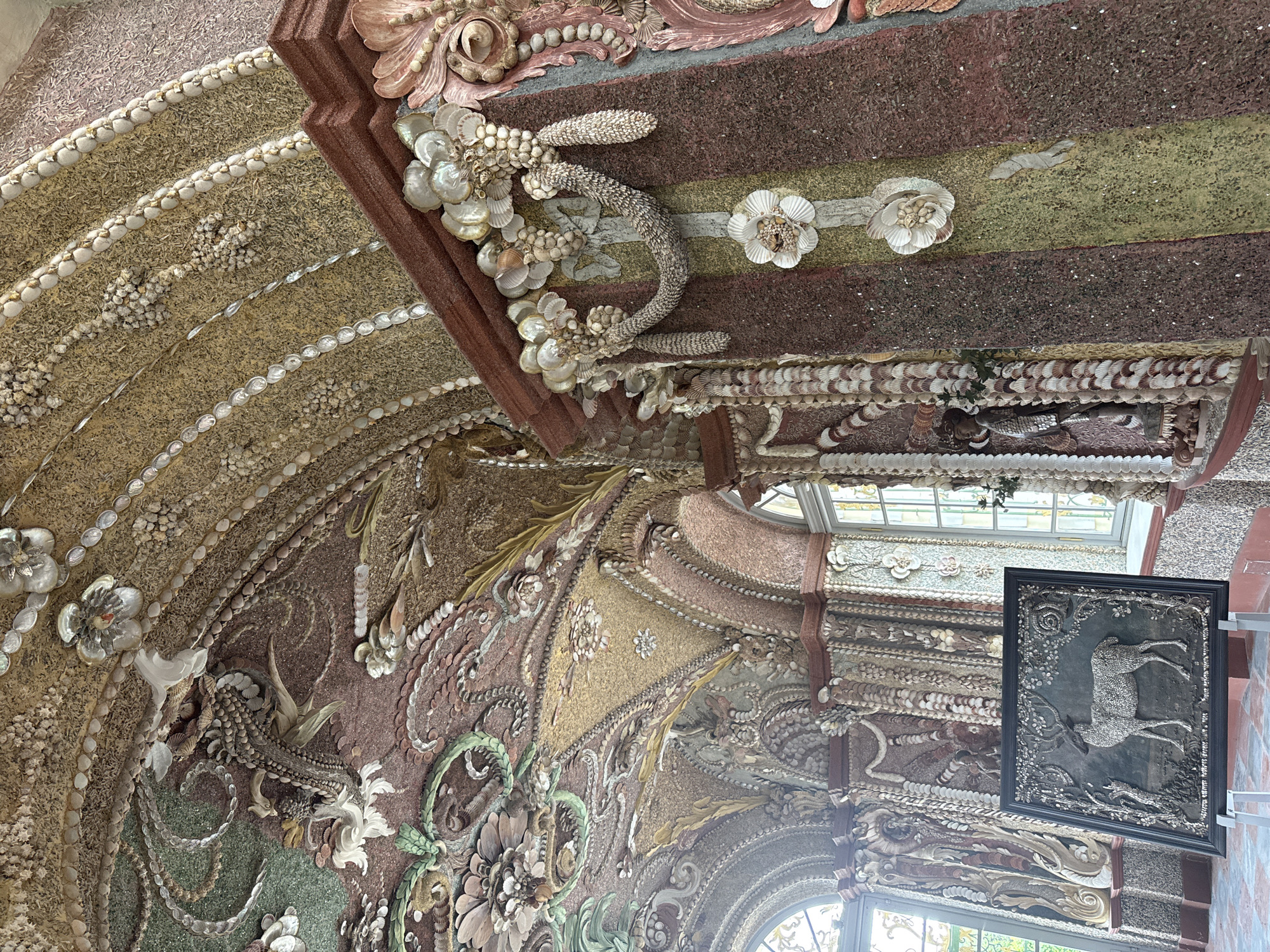The image captures an ornate interior with walls and ceilings elaborately decorated with seashells and various textured materials. The scene appears to be a part of a historical building in Moscow, Russia. The ceiling features multiple layers of decorative elements, including floral patterns, shell borders, and contrasting textures. A framed artwork depicting a running deer hangs on the wall, reflecting light from nearby windows. The architectural details point to a location with rich historical and artistic significance.