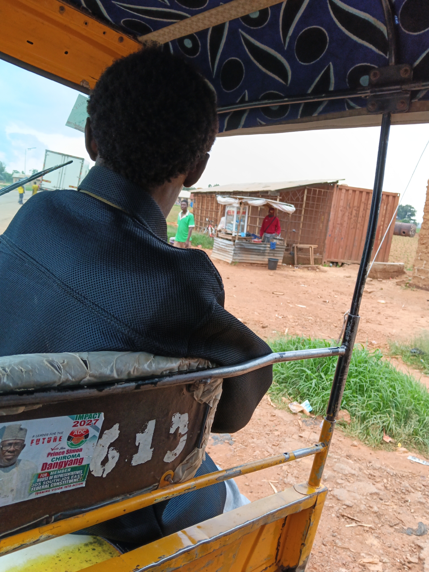 The image shows a person sitting in the passenger seat of a yellow auto-rickshaw, viewed from behind. The passenger is wearing a black jacket with a textured pattern. The auto-rickshaw has a blue and white patterned canopy overhead. In the background, there is a roadside stall with a person in a red shirt and another person in a green shirt. A large shipping container is also visible. The ground is dirt and there is green grass to the right. A sticker on the side of the auto-rickshaw features a picture of a man in a hat and text that reads "IMPACT 2027" and "FUTURE", along with other campaign-related slogans. The numbers "613" are painted on the side of the auto-rickshaw. The overall environment suggests a rural or semi-urban setting.
