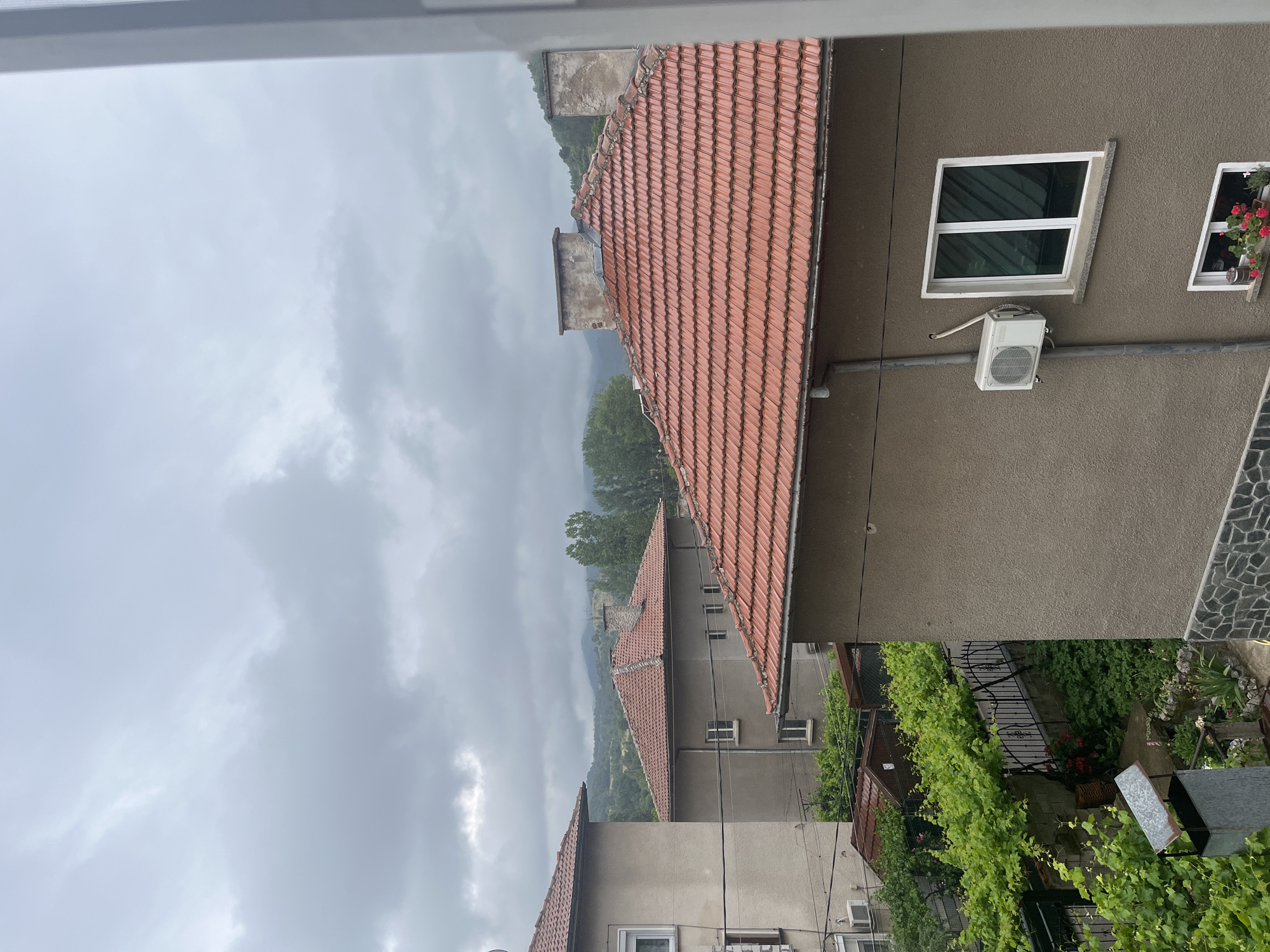 The image presents an overcast daytime view of a residential area, consistent with the provided context of Pokrovnik, Bulgaria. The perspective is from a window or balcony, looking down onto several buildings and lush greenery.
The sky is heavily clouded with various shades of grey, indicating an overcast day, possibly after or during light rain, as some of the terracotta-tiled roofs appear slightly damp. In the immediate foreground and to the right, a building with a textured, brownish-grey stucco facade is prominent. It features a modern white-framed window with dark panes, below which an outdoor air conditioning unit is mounted. Further right on the same wall, another white-framed window is adorned with flower boxes containing vibrant red blossoms, likely geraniums. A section of a wall finished with irregular stone tiles is also visible at the bottom right.
The mid-ground is characterized by multiple traditional red-tiled roofs, sloping downwards from right to left, indicating the terrain or architectural layout. Several chimneys rise from these roofs. Extensive green vegetation, including dense vines growing on trellises or fences and various trees, is abundant throughout the scene, particularly in the lower left and central areas, partially obscuring the lower levels of some buildings.
In the distant background, hazy, forested hills or mountains emerge beneath the cloudy sky, suggesting a natural, possibly mountainous, landscape surrounding the settlement. There are no people visible, and no discernible text appears in the image. The scene conveys a quiet, tranquil atmosphere in a rural or semi-rural European setting on a cloudy day.