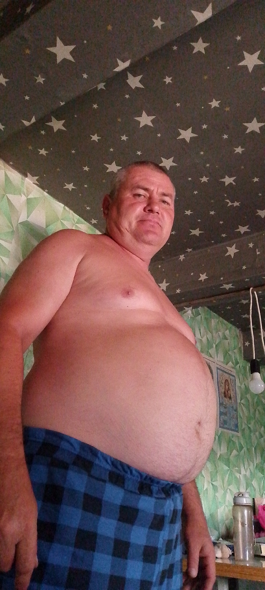 A low-angle, medium shot shows a shirtless, overweight man from the chest down, wearing blue and black plaid pajama bottoms. The man has fair skin, a rounded stomach, and a visible chest. He is looking directly at the camera with a neutral expression. The background features a gray ceiling covered in white and yellow stars, and a green patterned wallpaper with a framed image of a religious figure. A lightbulb hangs from the ceiling on the right side of the frame, and a water bottle and other items are visible on a surface to the right of the man. The scene appears to be indoors, likely in a bedroom or living area.