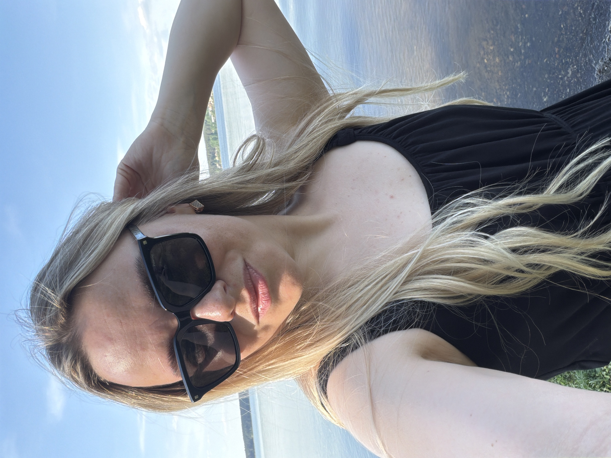 A woman with long blonde hair and sunglasses takes a selfie outdoors. She is wearing a black top. The background includes water and trees, suggesting a lakeside or riverside location. It is daytime with clear sky. The location is Votkinsk, Russia, although the specific spot is unidentifiable without further landmarks or information.
