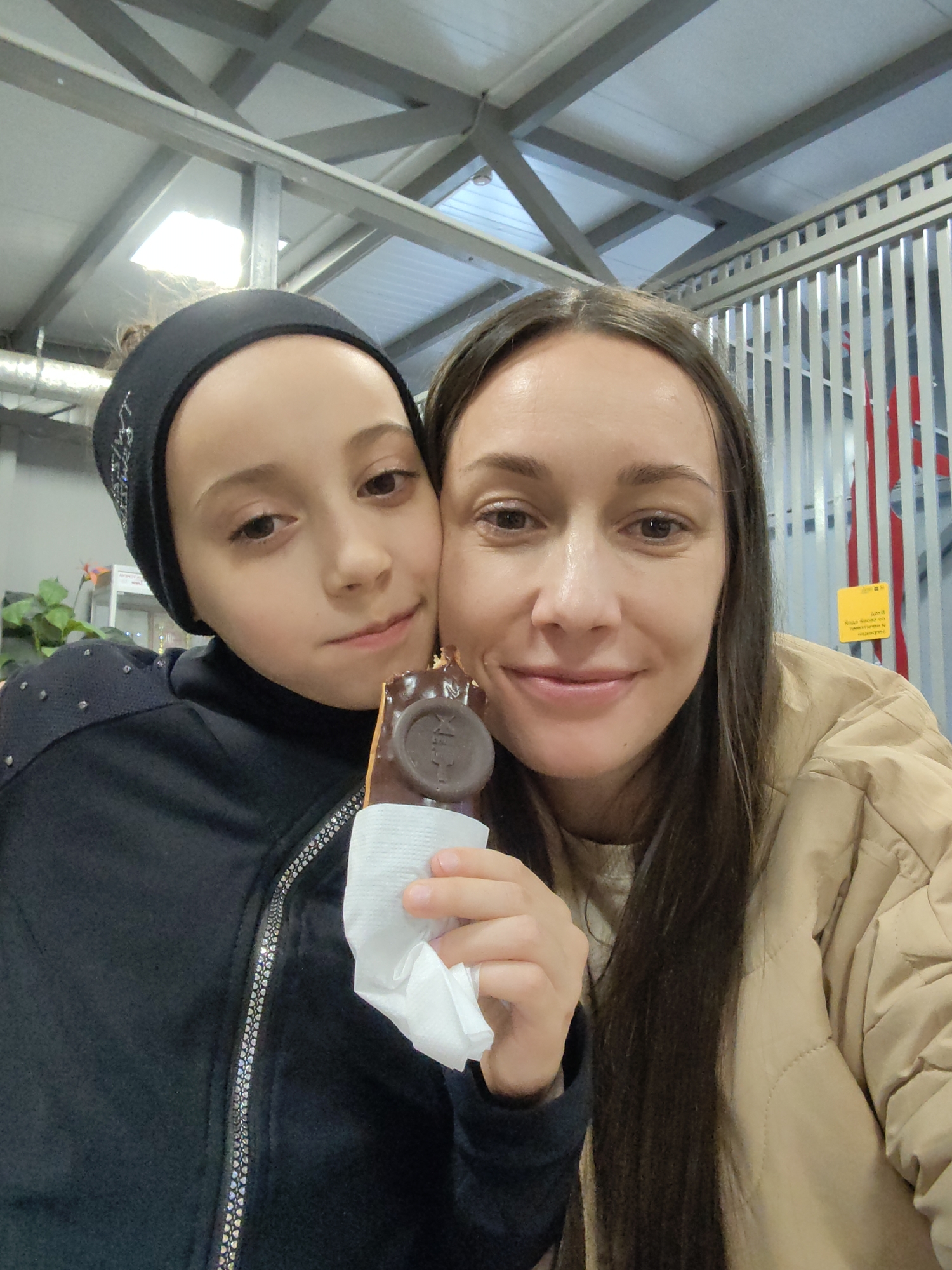 A selfie captures a woman and a girl, holding an ice cream bar, likely in Vidnoye, Russia. The girl, positioned to the left, wears a black head band and a jacket with metallic accents. The woman, on the right, wears a tan quilted jacket. They stand against a background of silver vertical bars. The ice cream bar is held in a napkin. The ceiling is white with silver beams.