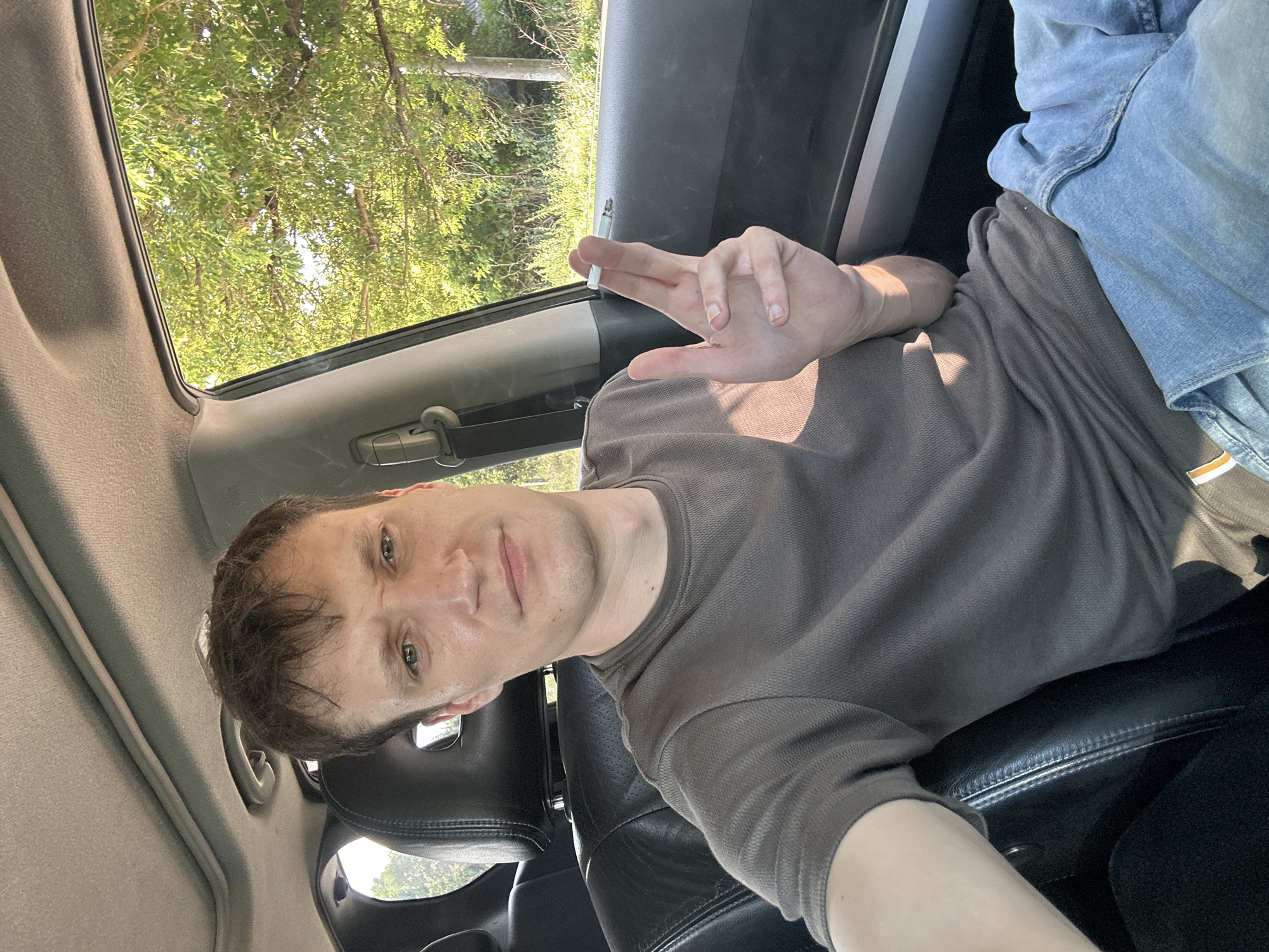 A person is seated in the driver's seat of a car, holding a cigarette between their index and middle fingers, with the cigarette lit and smoldering. The person is wearing a dark brown t-shirt and blue jeans. The car's interior is visible, including the leather seats and the car's headliner. Sunlight streams through the car's window, illuminating the person and the interior. Outside the window, trees and foliage are visible, suggesting an outdoor setting, possibly in a rural area or a park. The scene does not contain any identifiable landmarks or elements that would specifically place it in the city of Makiv, Ukraine.