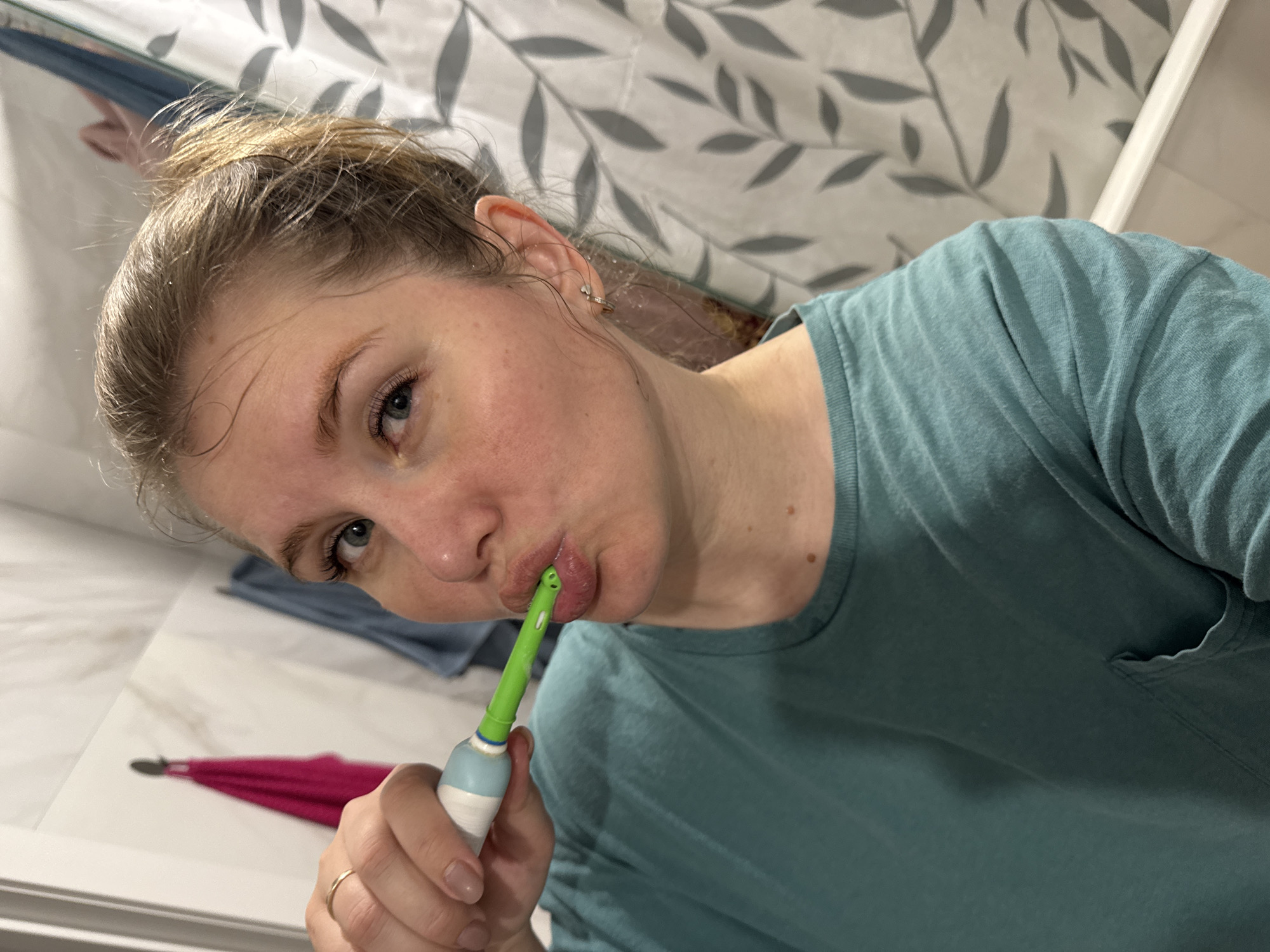A woman is brushing her teeth in a bathroom. She is wearing a turquoise t-shirt and a gold ring on her left ring finger. Her hair is up in a bun. She is holding an electric toothbrush with a green brush head. The bathroom has a shower curtain with a leaf pattern, a white marble wall, and a pink towel. The location is not identifiable as Ферма, Russia.
