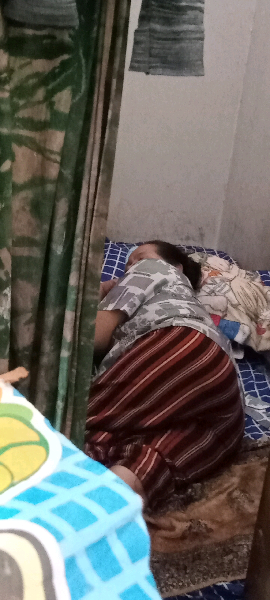 An overhead, medium shot shows a person lying on a bed, asleep. The person is wearing a grey and white patterned t-shirt and a red and white striped skirt. They are lying on their side, with their head resting on a pillow. The bed is covered in a blue and white patterned sheet, and a blanket with a floral pattern. A green curtain hangs to the left of the bed. The wall behind the bed is white and slightly textured. The scene appears to be inside a room in Delanggu, Indonesia.
