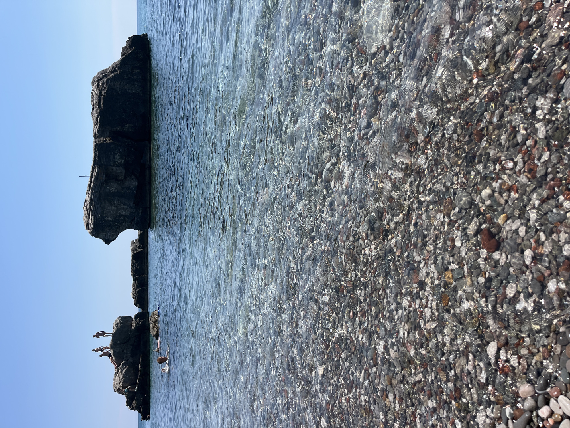 The image depicts a sunny day at a rocky beach in Zagrađe, Montenegro. The scene is split between a clear blue sky and the turquoise water of the sea, which laps against a shore covered in smooth, colorful pebbles. In the distance, large, dark rock formations rise out of the water, some of which are accessible by swimmers.
Several people are visible, enjoying the refreshing water and the opportunity for cliff diving. On one of the rock formations, three individuals are in the process of jumping into the sea, captured mid-air. Another person is in the water, swimming near the rocks. The clear water allows for a view of the pebbly seabed. The overall atmosphere is one of leisure and fun, characteristic of a summer day at the beach. The time of day is likely midday, given the bright sunlight and clear skies. There is no visible text in the image.
