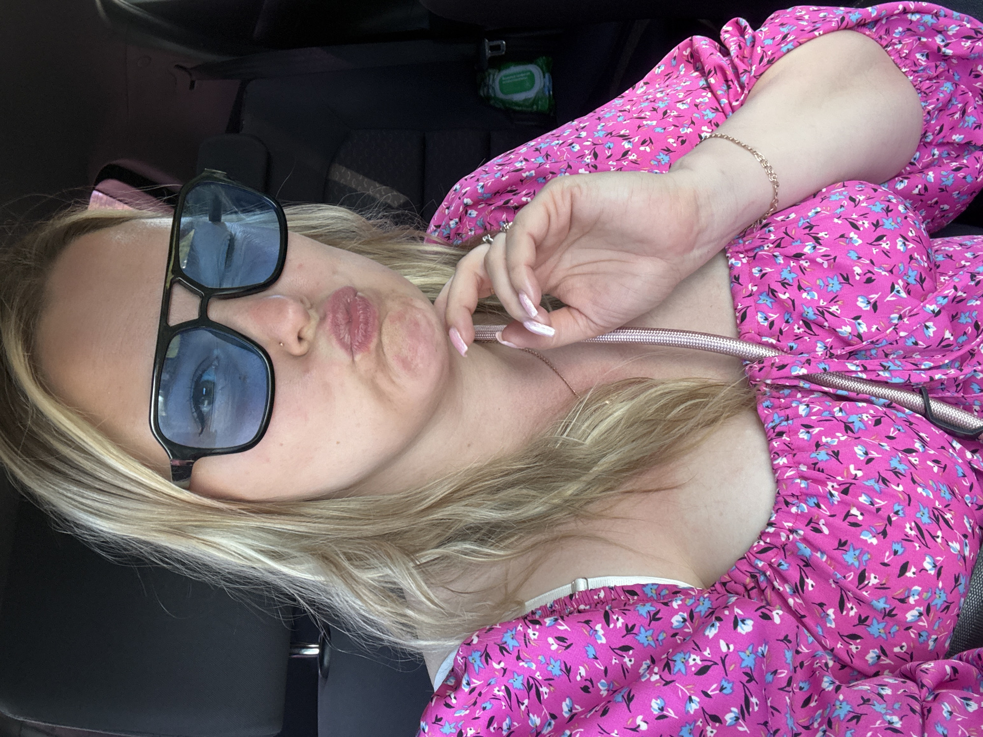 A woman is taking a selfie inside a vehicle, likely a car. She is wearing large black-rimmed sunglasses and a pink, floral-patterned dress with a low neckline. Her blonde hair is parted and falls around her face. She is holding her hand up near her face, fingers slightly curled. She is puckering her lips as part of the selfie expression. There are car seats and some green wipes visible in the background. There is no indication of Noviy, Russia in this image.