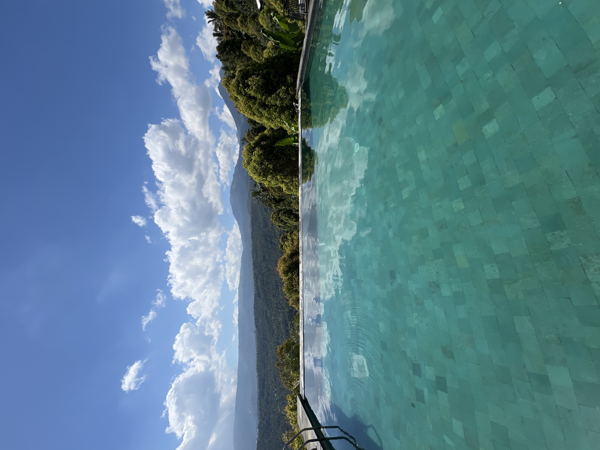An infinity swimming pool with turquoise tiles reflects the sky and clouds. In the background, lush green hills and mountains are visible under a bright blue sky with scattered white clouds. The scene appears to be a resort or private residence in a tropical location. The location is Busungbiu, Indonesia.