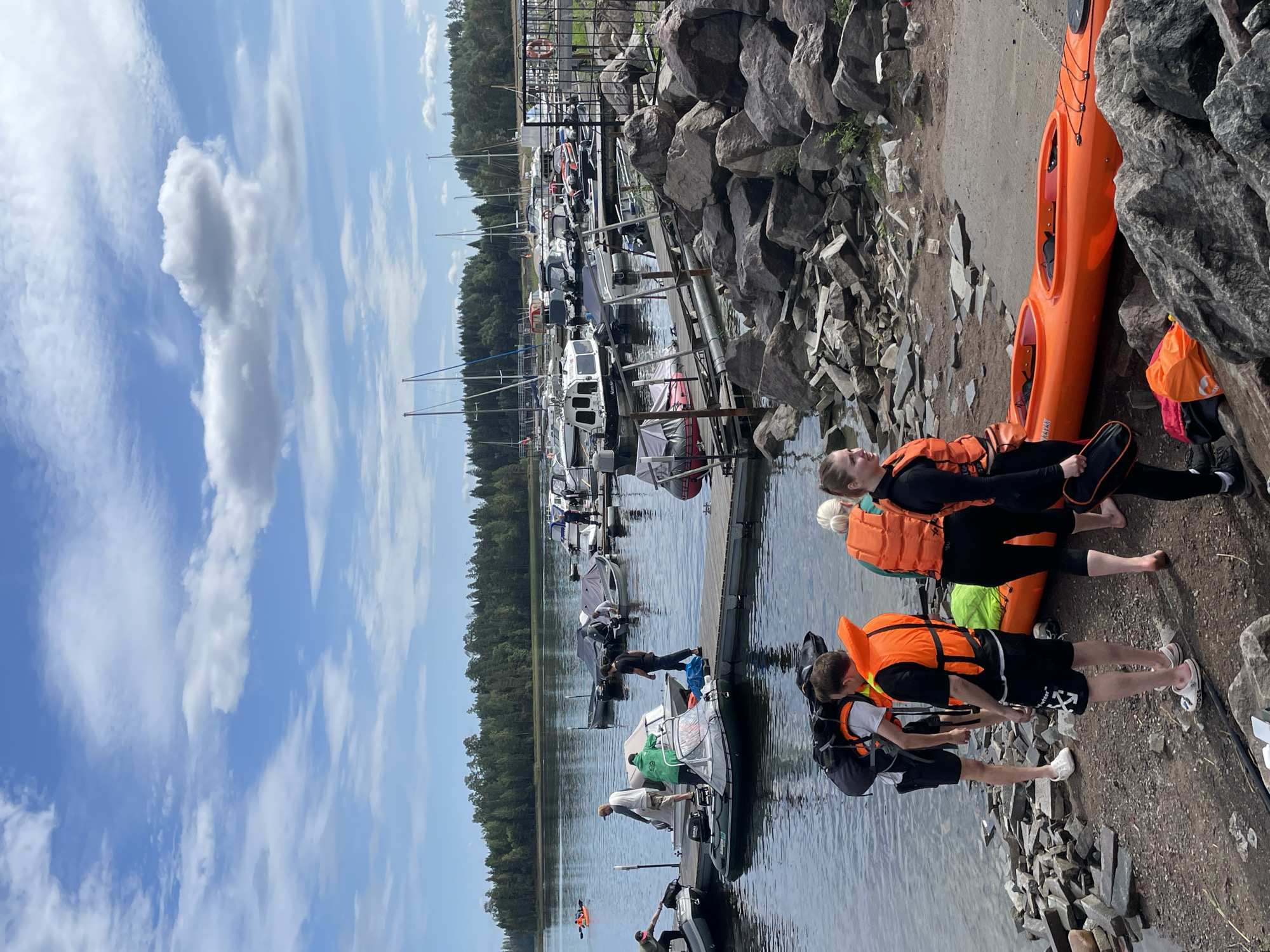 The image depicts a waterside scene with a marina and a rocky shoreline. In the foreground, several individuals are gathered near an orange kayak. Two people are wearing orange life vests, one of whom has a large backpack. Another person, a woman, is dressed in dark clothing and an orange life vest, holding a bag. A large orange kayak lies on the ground beside them.
The midground shows a wooden pier extending into the water, with several boats docked. Some of these are small motorboats, while others appear to be sailboats in the distance. People are seen interacting with the boats on the pier.
The background features a dense forest of green trees, suggesting a natural, perhaps rural, environment. The sky is blue with scattered white clouds. The overall impression is a recreational area, likely a marina or a lakefront park, where people are preparing for or have just finished water activities. The location is identified as Berozovo, Russia.