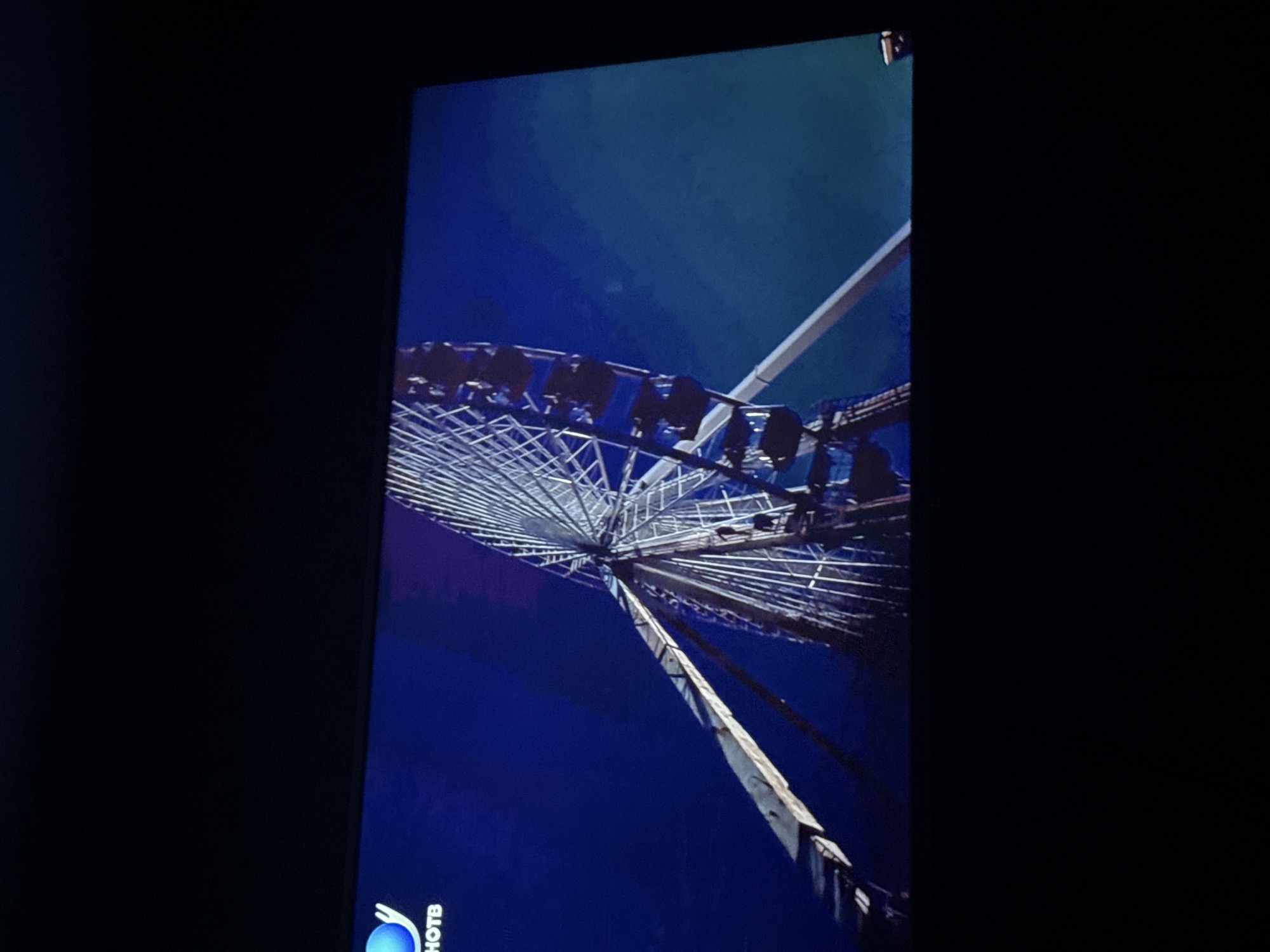 A view of a Ferris wheel is seen from the ground, looking up. The wheel is metallic and geometric, against a dark blue sky. Individual gondolas hang from the wheel's rim. A structural support beam extends upwards. The image is displayed on a television screen, as evidenced by the surrounding black frame and the word "PHOTOS" in the lower left corner. There is no evidence in the picture that this image is in the city of Mytishchi, Russia.