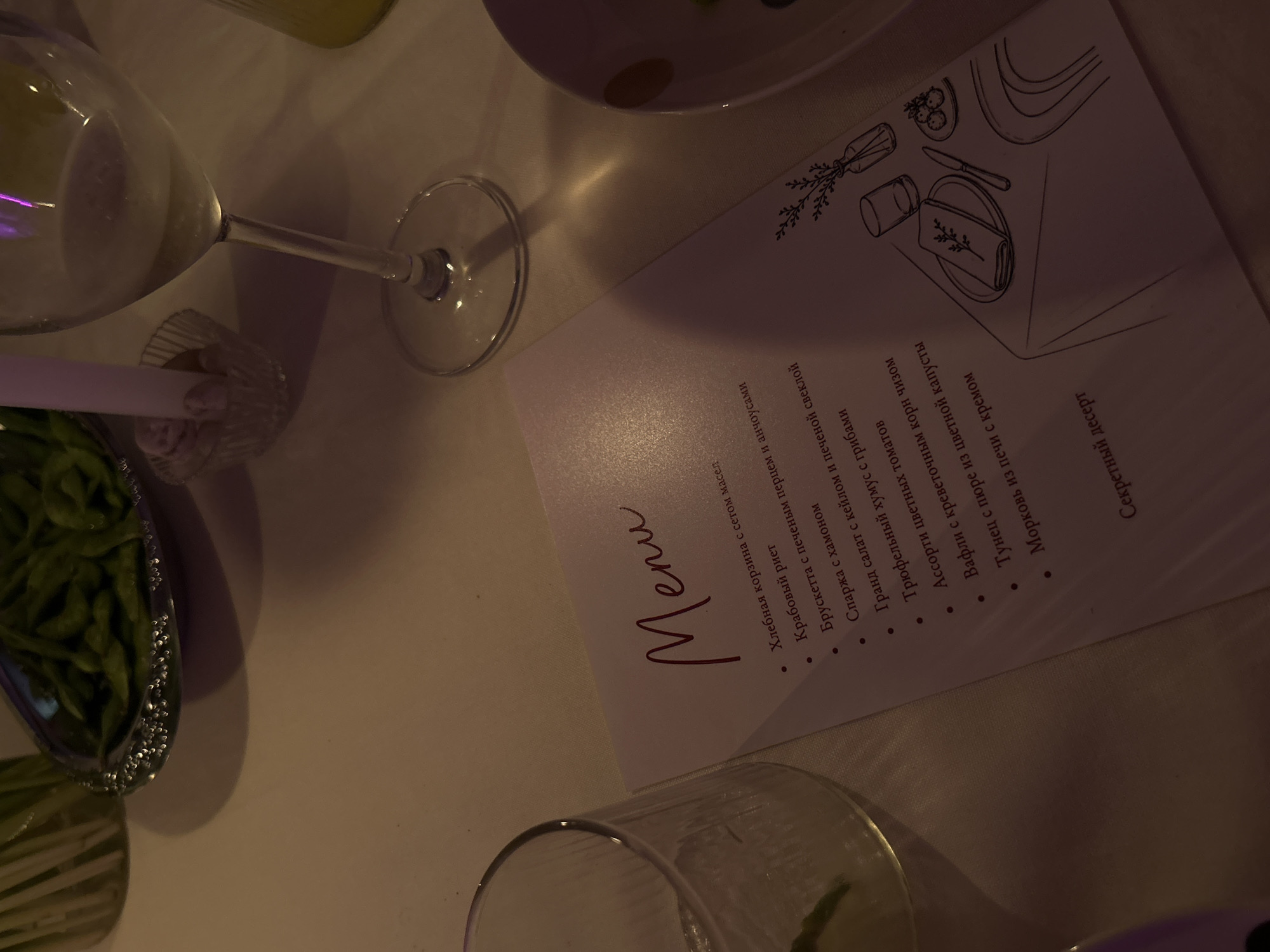 A dimly lit indoor scene features a white menu on a table. The menu has a handwritten-style title "Menu" in burgundy and a bulleted list of food items. Some of the visible items on the menu include "Хлебная корзина с сетом масел," "Крабовый риет," "Брускетта с печеным перцем и анчоусами," "Спаржa с хамоном," "Гранд салат с кейлом и печеной свеклой," "Трюфельный хумус с грибами," "Ассорти цветных томатов," "Вафли с креветочнм корни чизоm," "Тунец с пюре из цветной капусты," and "Морковь из печи с кремом." At the bottom of the menu, the text "Секретный десерт" is visible. To the right of the menu, a black line drawing depicts a table setting with a plate, napkin, knife, and small round food items. Further to the right, a sketch of a vase with branches and a glass is also present. The background shows a glass of white wine, a glass with ice and lime, and a dish of green edamame beans. The environment appears to be a restaurant setting, possibly in Chang Phueak, Thailand, given the provided context. There are no people visible in the image.