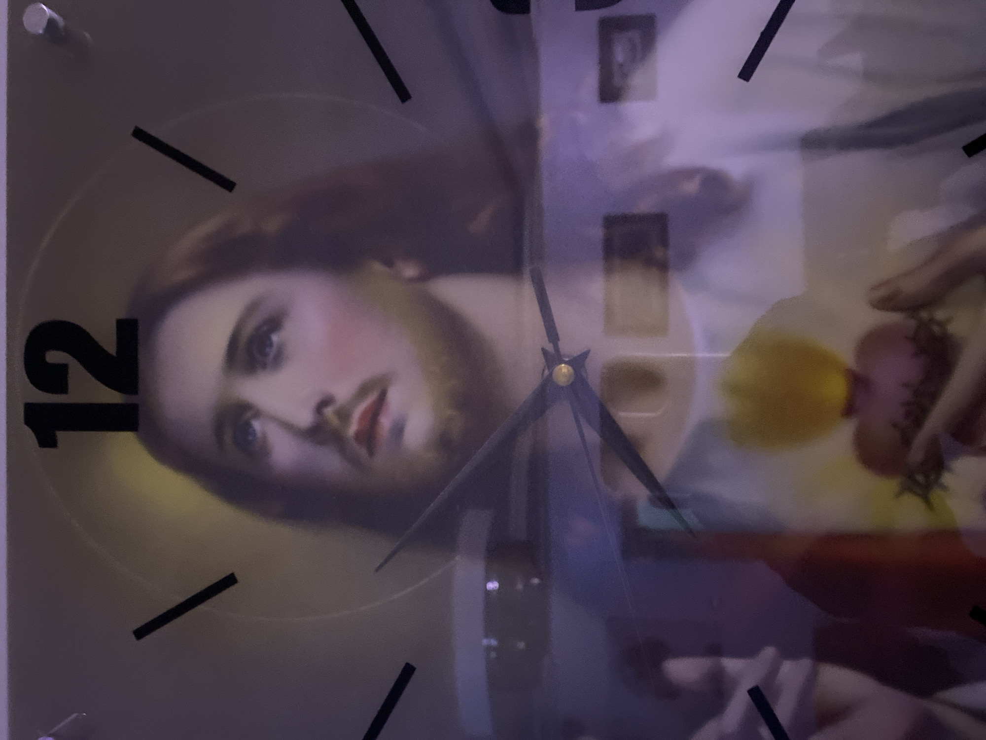 The image shows a close-up of a wall clock with a depiction of Jesus Christ. The clock face features Roman numerals and has visible hour and minute hands. The image of Jesus is centered on the clock face, with his head and upper torso visible. The background of the clock is a light color with a subtle halo effect around Jesus' head. The clock appears to be made of glass or a similar reflective material, as there are some reflections visible on the surface. The image is taken from a frontal perspective, slightly angled. The quality of the image is somewhat grainy, suggesting it might have been taken in low light conditions or with a lower resolution camera. There is no information within the image or the provided crops to indicate a location in Giginyu, Nigeria.