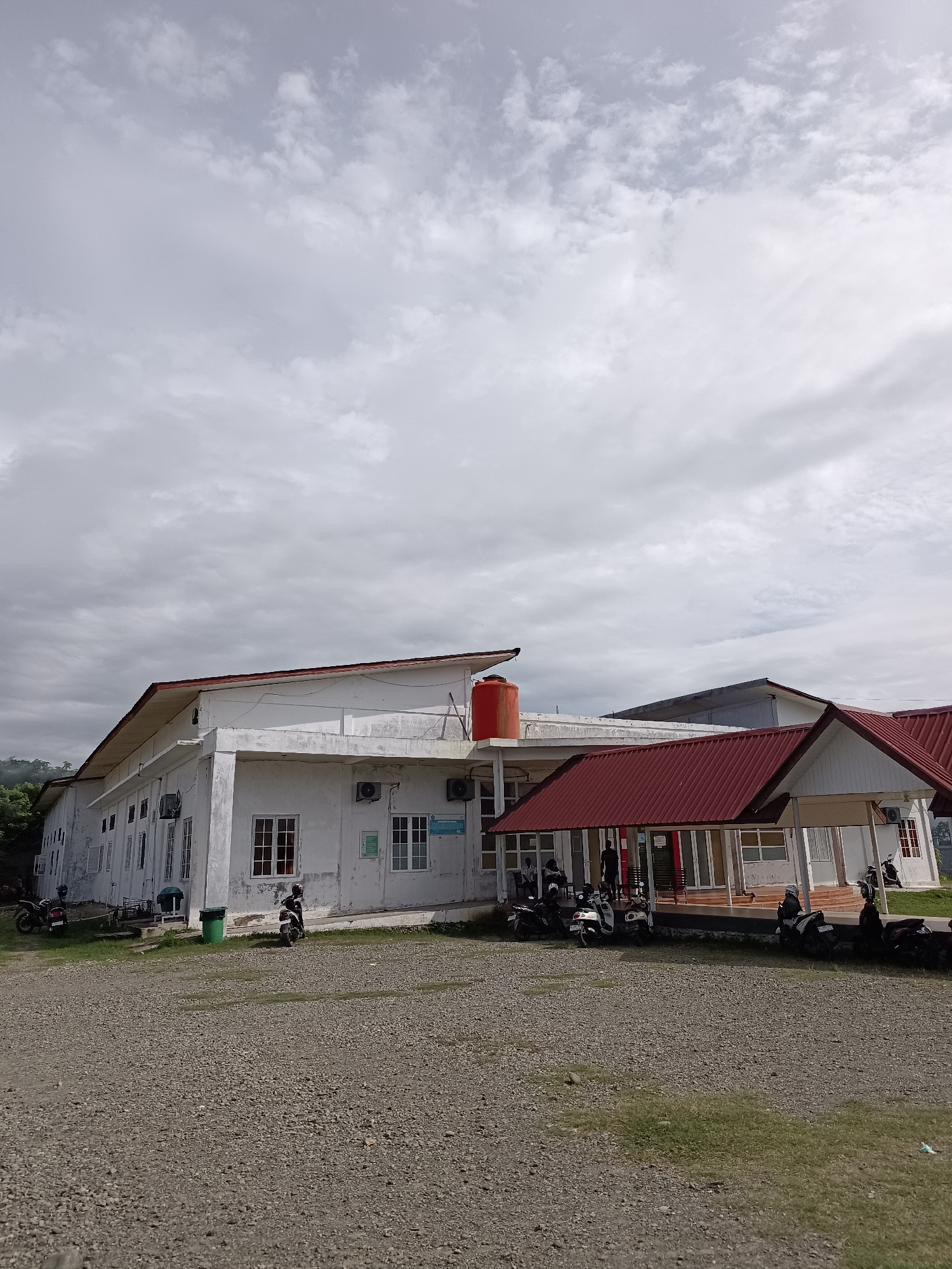 The image depicts a white, two-story building with a red-tiled roof, situated in a hilly, possibly rural, environment. The building appears to be a public facility, possibly a school or administrative office, with several windows and air conditioning units visible on its exterior. In front of the building, there is a gravel-covered parking area with several motorcycles parked. Some people are visible near the entrance of the building, standing or walking. The sky is overcast with clouds, suggesting a potentially cloudy or rainy day. In the background, lush green trees and hills are visible, indicating a natural, undeveloped landscape. The overall impression is that of a functional building in a serene, natural setting, likely within or near the city of Mesjid Reuntoh, Indonesia.