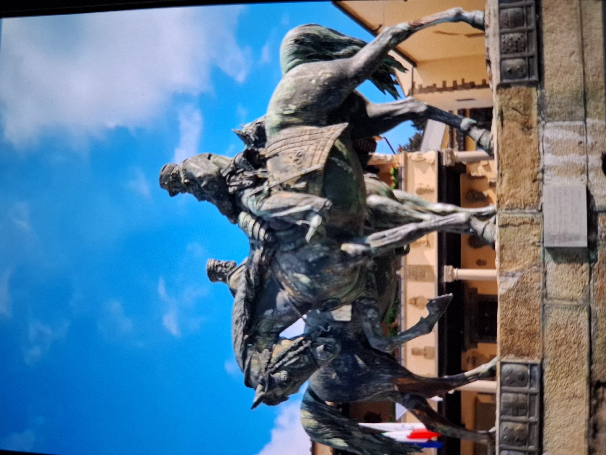 A bronze equestrian statue featuring two figures on horseback stands on a stone pedestal in an outdoor plaza. The statue depicts two men in historical attire, each riding a horse. The horses are rendered in a dynamic pose, appearing to be in motion. Behind the statue, a building with warm-toned walls and architectural columns is visible under a bright blue sky with scattered clouds. Flags, appearing to be red, white, and blue, are also visible to the left of the statue, attached to a pole. The statue is weathered, exhibiting a greenish patina characteristic of aged bronze. The stone pedestal is constructed from rough-hewn blocks, some of which are intricately carved.