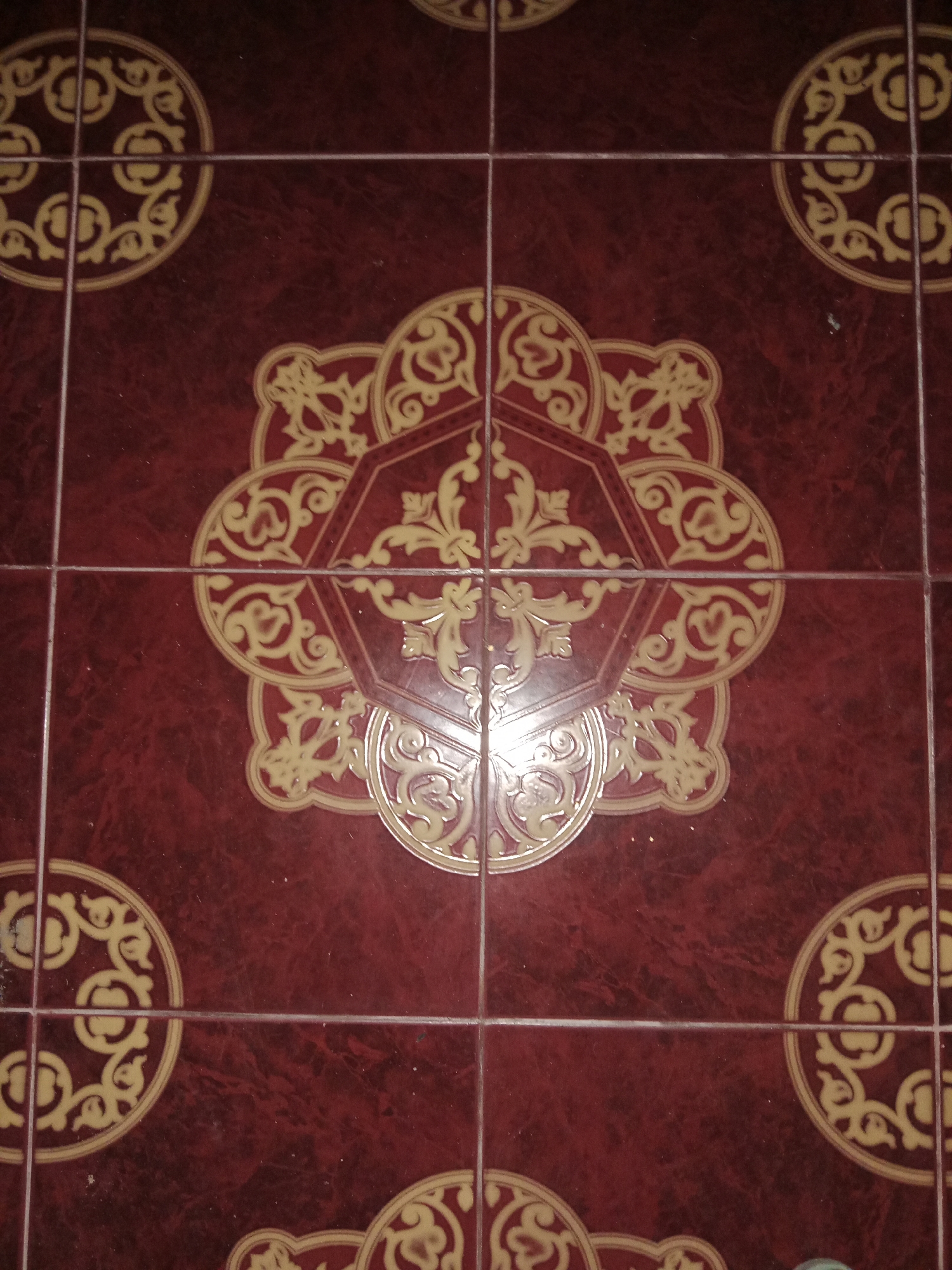 The image shows a close-up, overhead view of a tiled floor. The tiles are a deep, dark red color with a subtle marbling effect. Each tile is decorated with an ornate, golden-yellow pattern. In the center of the image, a larger, more complex golden design is visible, spanning across multiple tiles. This central design appears to be an octagonal shape with intricate floral or arabesque motifs. Smaller, circular golden patterns are also visible in the corners of the frame, appearing on individual tiles. The grout lines between the tiles are thin and white, creating a grid over the surface. The lighting suggests an indoor environment, possibly a home or public space. The image does not depict any people, actions, or interactions. The location in Ringinrejo, Indonesia is not visually evident from the image itself.