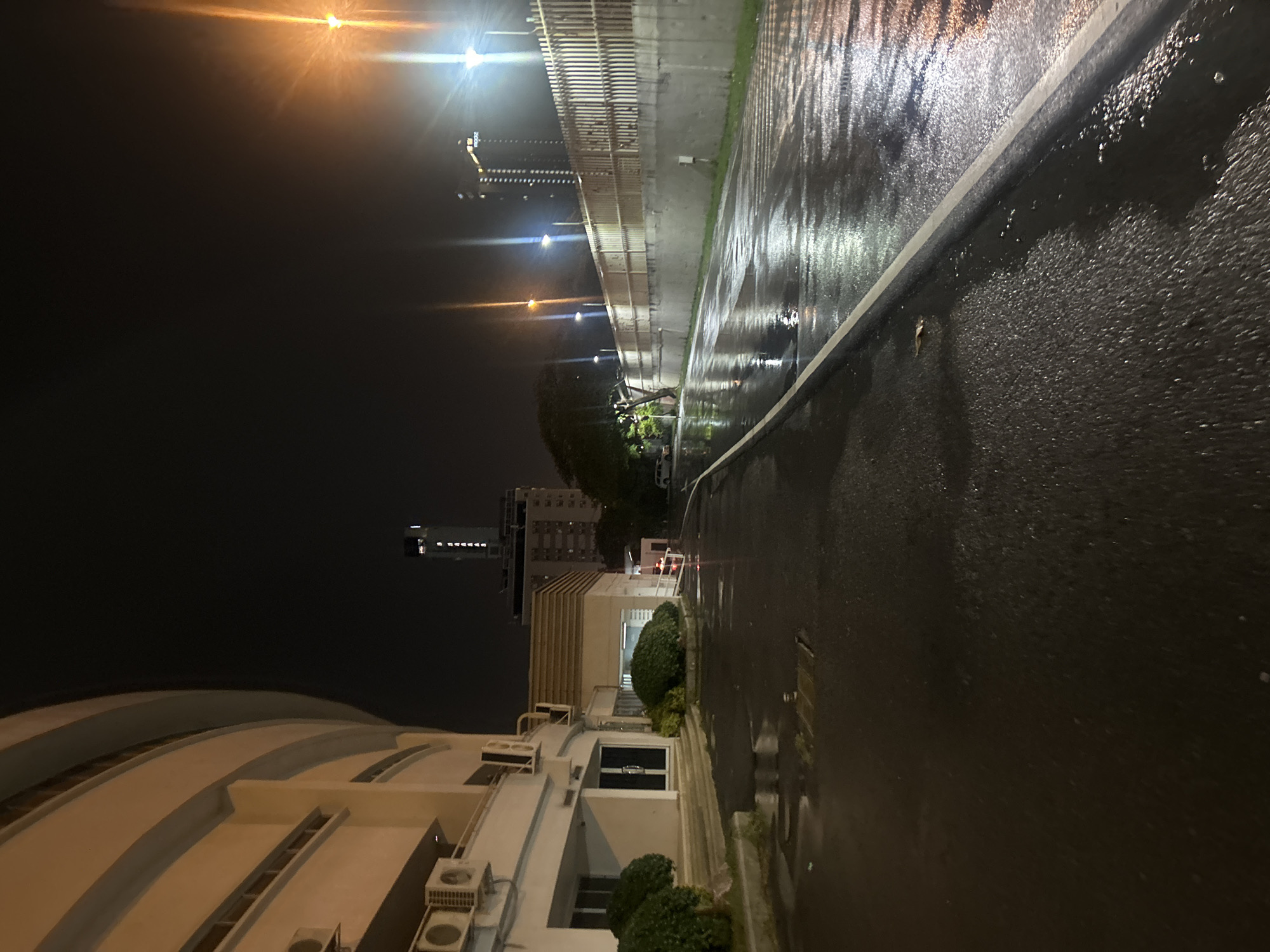 The image depicts a wet, paved area at night, likely a street or driveway, in Khlong Luang Town Municipality, Thailand. The ground is dark and reflective due to recent rain. To the left, a curved, modern building with pale walls and horizontal banding is visible, featuring air conditioning units mounted on its exterior. Some lush green bushes are present near the base of this building. To the right, a concrete wall topped with a slatted fence runs along the scene, illuminated by streetlights. Beyond the fence, a few trees are visible. In the background, several tall buildings are illuminated against the dark night sky, including one with the name "PARANAS" clearly visible in vertical white lettering. A few streetlights are also present, casting bright glows and reflections on the wet surface. The scene appears to be an urban or suburban environment after a rainfall.