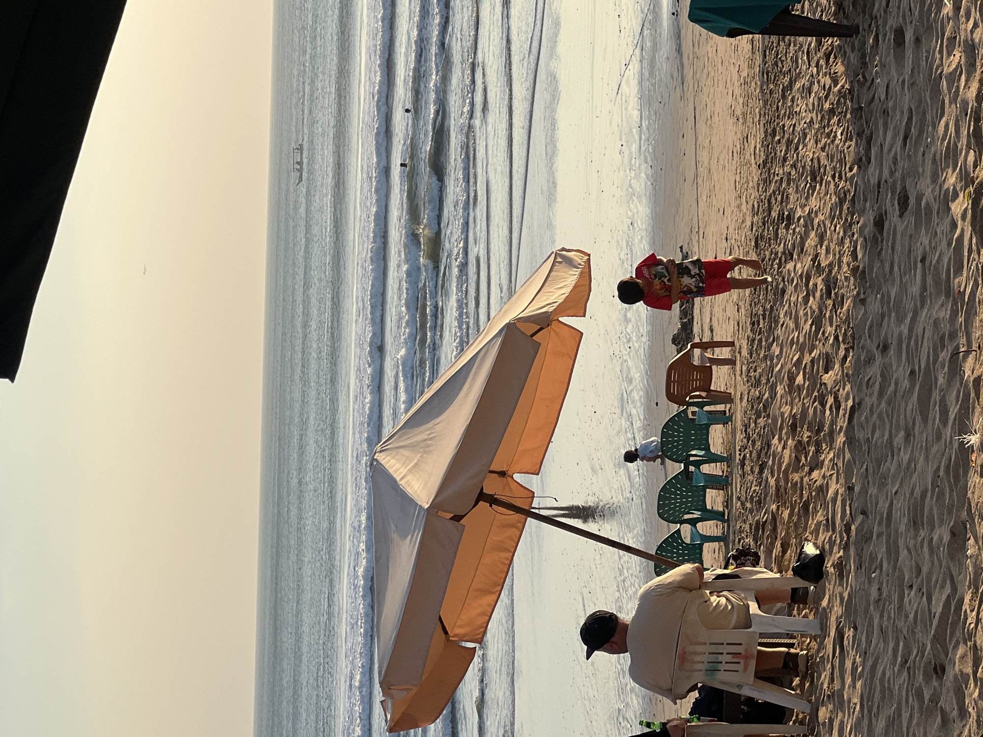 An overhead shot captures a beach scene in Kuta, Indonesia, with the ocean and sky dominating the background. In the foreground, sand is disturbed by footprints and the shadows of beach umbrellas. Several plastic chairs, some occupied, are arranged on the sand. A large, beige beach umbrella shades a portion of the sand where a person sits facing away from the camera, wearing a light-colored t-shirt and a black cap. To the right, a young boy in a red t-shirt and red shorts walks across the sand. In the middle ground, several green plastic chairs are visible, and further back, a child crouches near the water's edge. The ocean has moderate waves, and several people are visible swimming or wading in the water. A small boat and a distant airplane are present in the hazy sky. The overall lighting suggests it is late afternoon or early evening, with a warm glow over the scene.