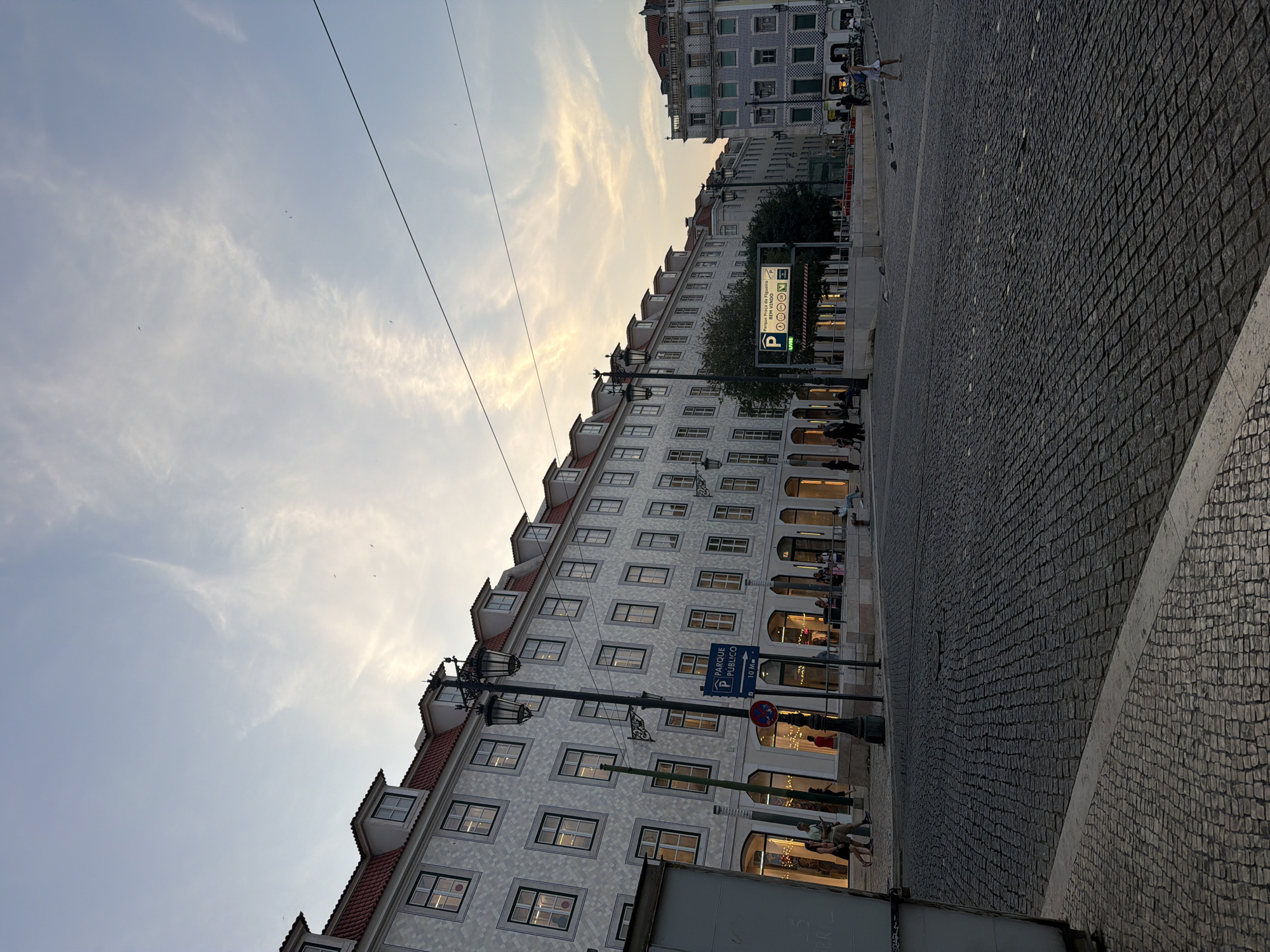 This is a medium-shot, outdoor, eye-level photograph of a cobblestone street in Lisbon, Portugal, during twilight. The sky is a pale blue with wisps of clouds, and the sun appears to have recently set, casting a warm glow on the horizon.
Dominating the left side of the frame is a long, multi-story building with a red tiled roof and dormer windows. The facade is decorated with a repetitive, light-colored diamond pattern. The ground floor features large storefront windows, some illuminated from within, revealing displays and people inside. A black lamppost with two ornate lamps stands prominently in front of this building.
To the right, the street opens into a plaza. A digital parking sign with Portuguese text ("PARQUE PÚBLICO") and other symbols is visible, indicating a public parking area. Further in the background, another building with a blue and white tiled facade is visible, as well as trees and more streetlights.
Several people are present in the scene. A few individuals are walking on the sidewalk in front of the storefronts, and others are sitting on a low wall. One person is walking across the cobblestone plaza in the foreground. The cobblestone pavement itself is a prominent feature, laid out in a dense, irregular pattern. The overall atmosphere is calm and urban, with the ambient light suggesting the transition from day to night.
